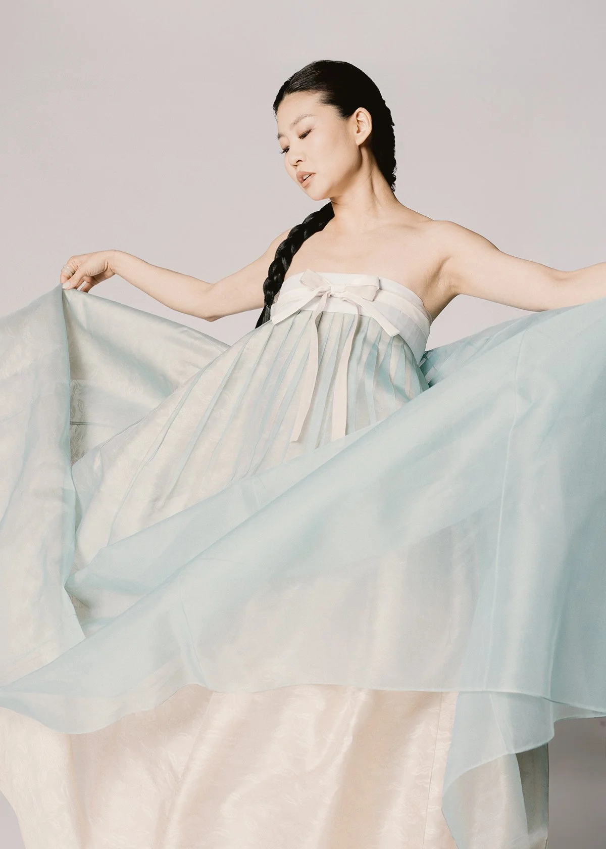 Woman with long black hair in braid wearing a strapless light-colored dress, holding out the fabric in a flowing pose against a plain background.