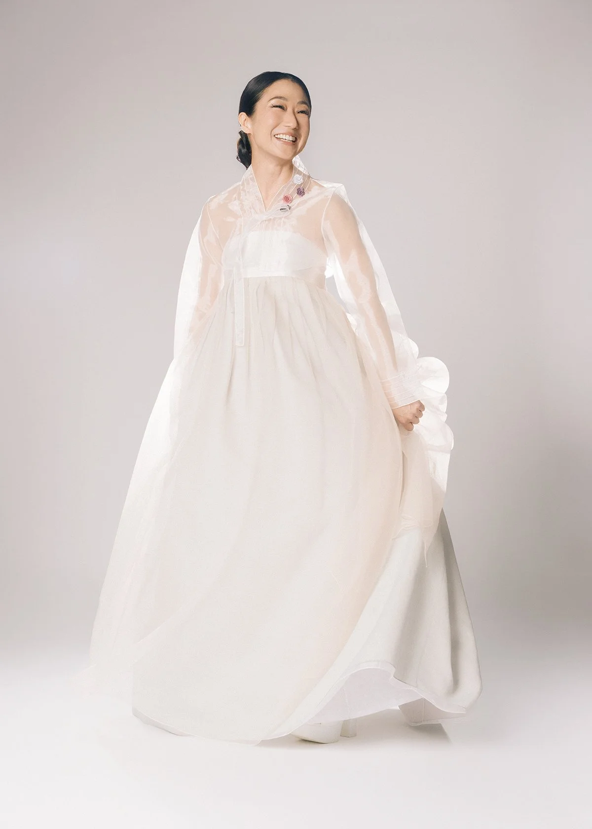 A woman smiling in a traditional Korean hanbok with a white jeogori and cream-colored chima, standing against a plain light background.