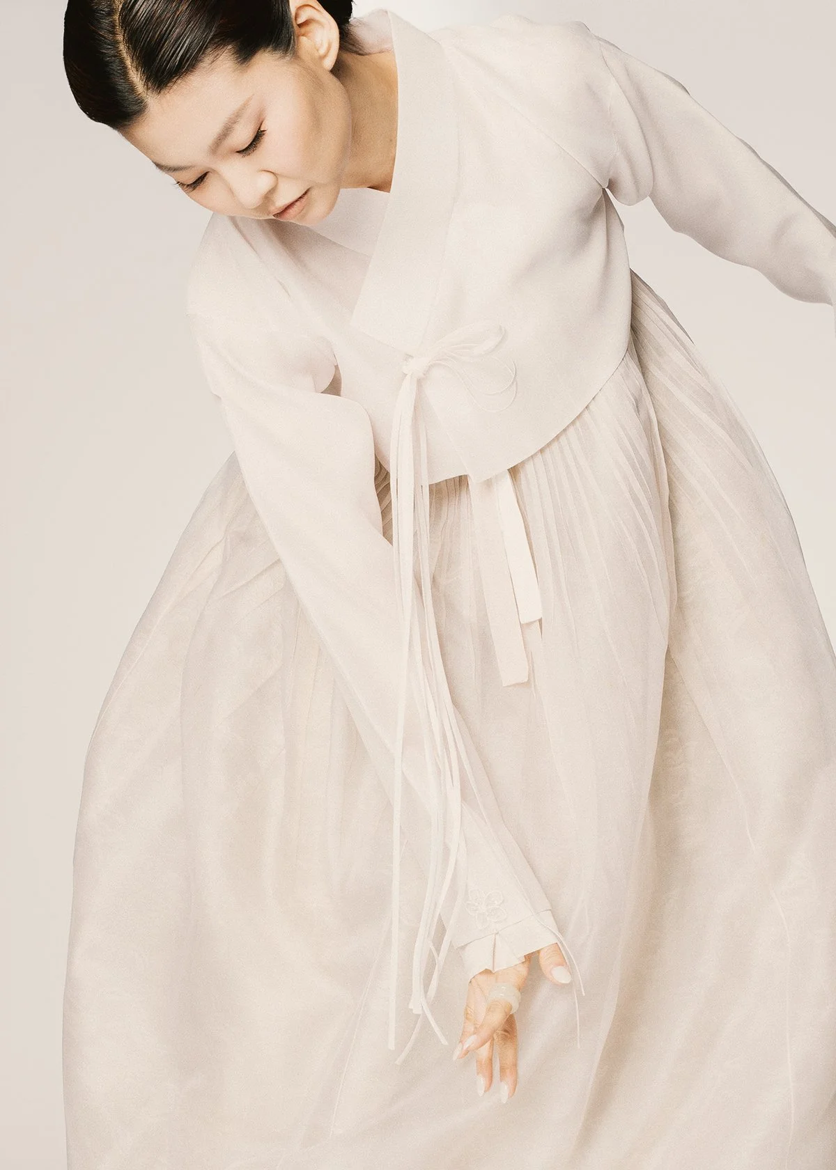 Close-up of a woman in traditional Korean clothing, Hanbok, with a white jeogori jacket and a flowing, pleated skirt, against a neutral background.