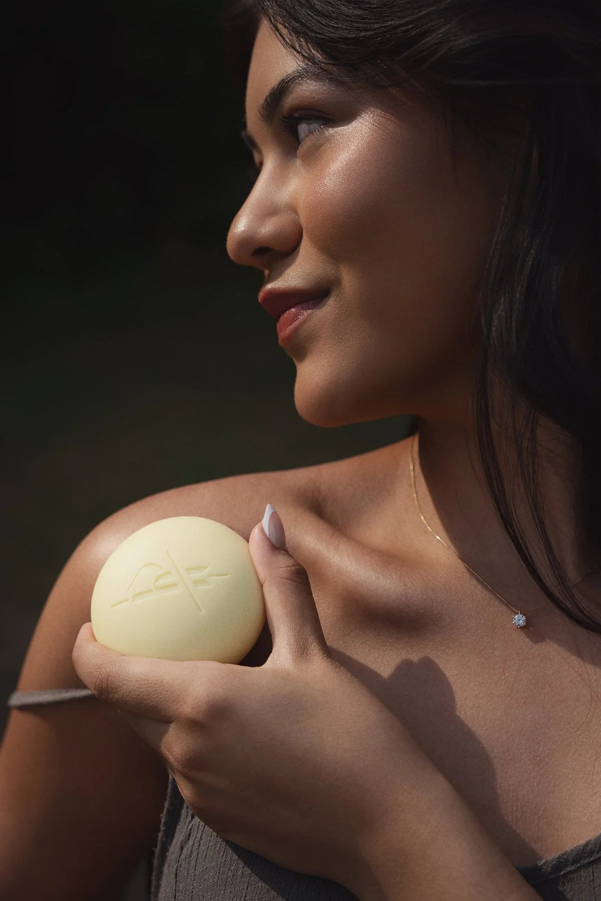 A woman with dark hair and a necklace holding a yellow soap bar with a logo, looking away with a gentle smile.