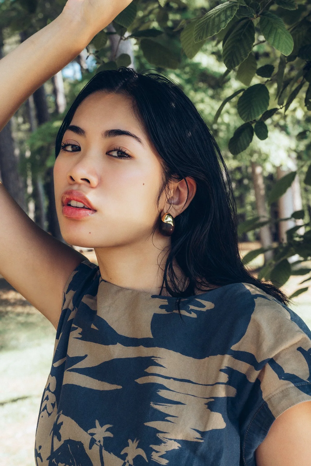 A woman with long dark hair and earrings poses outdoors amid greenery, wearing a camouflage patterned shirt.