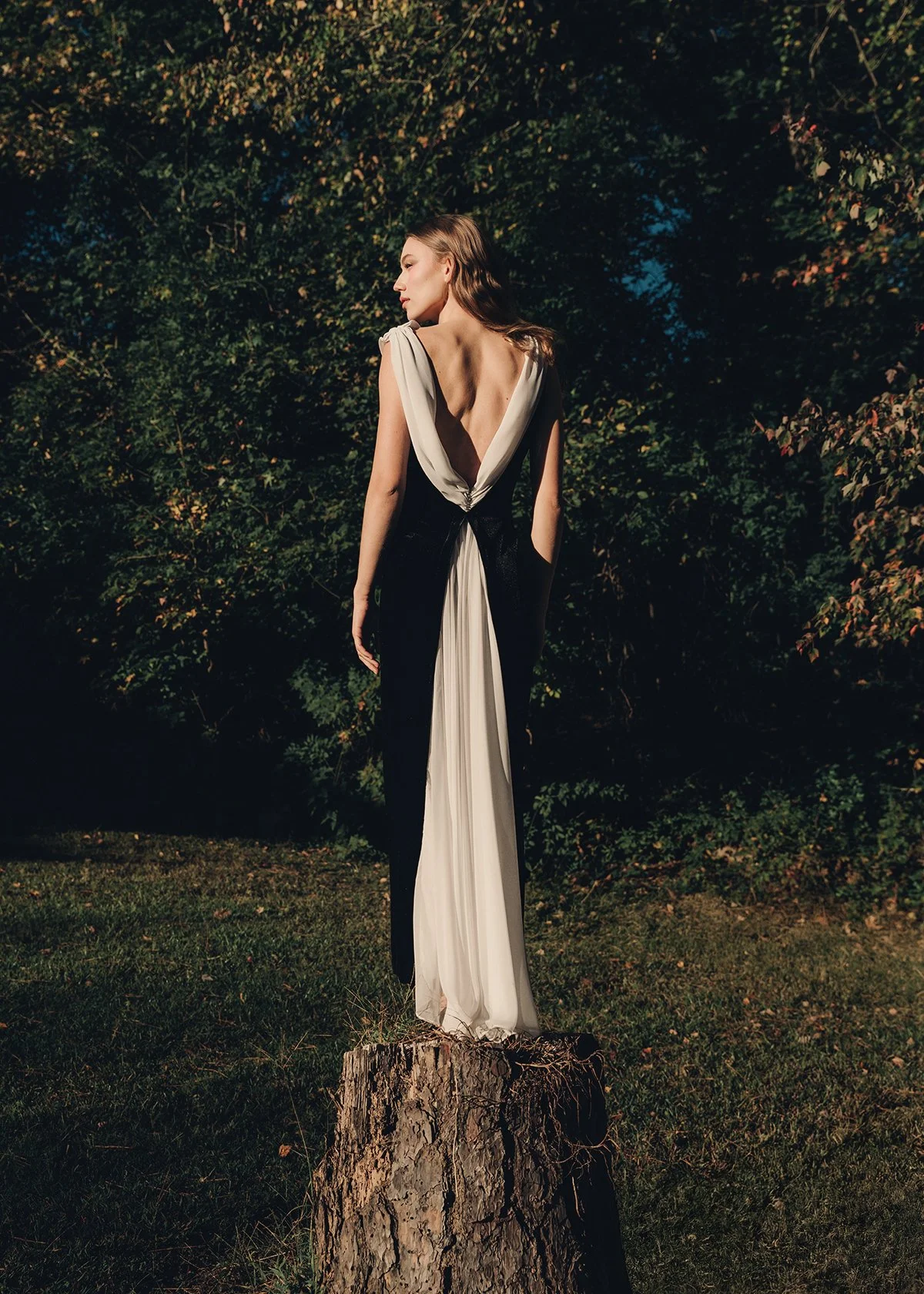 A woman in a black and white elegant dress standing on a tree stump outdoors at dusk, with dense green foliage in the background.