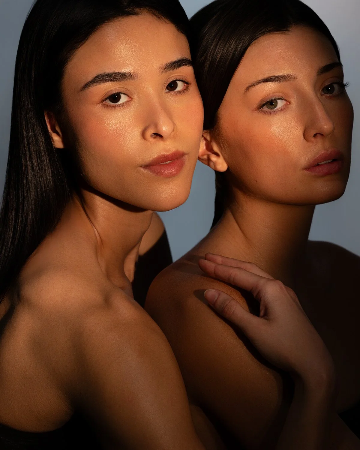 Two women with long dark hair and minimal makeup pose closely together against a neutral background, with one resting her hand on the other's shoulder.