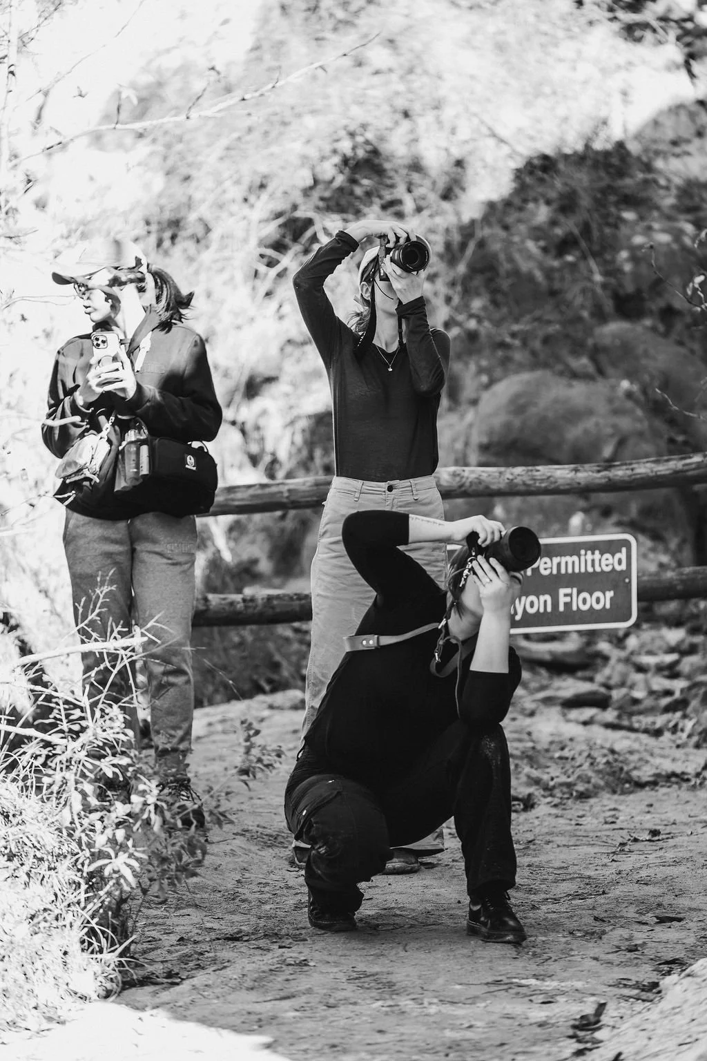 Three women taking photos with cameras in a wooded outdoor area, one squatting, one standing, and one looking at her phone while holding a camera. There is a sign in the background that reads 'Permitted Canyon Floor'.