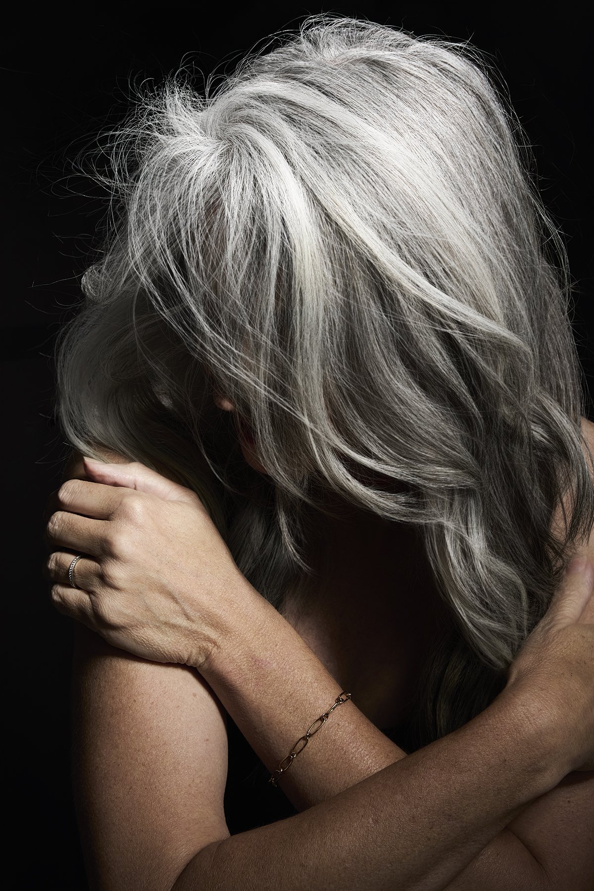 A woman with silver hair covering her face, hugging herself against a black background.