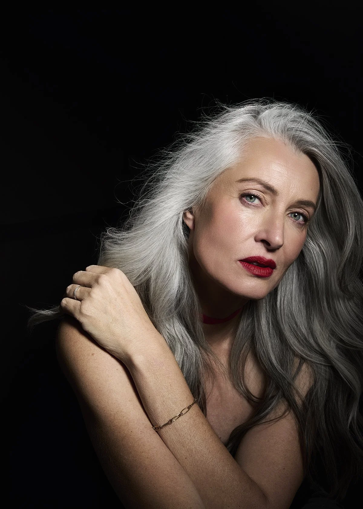 A portrait of a woman with long gray hair, blue eyes, and red lipstick, posing against a black background.