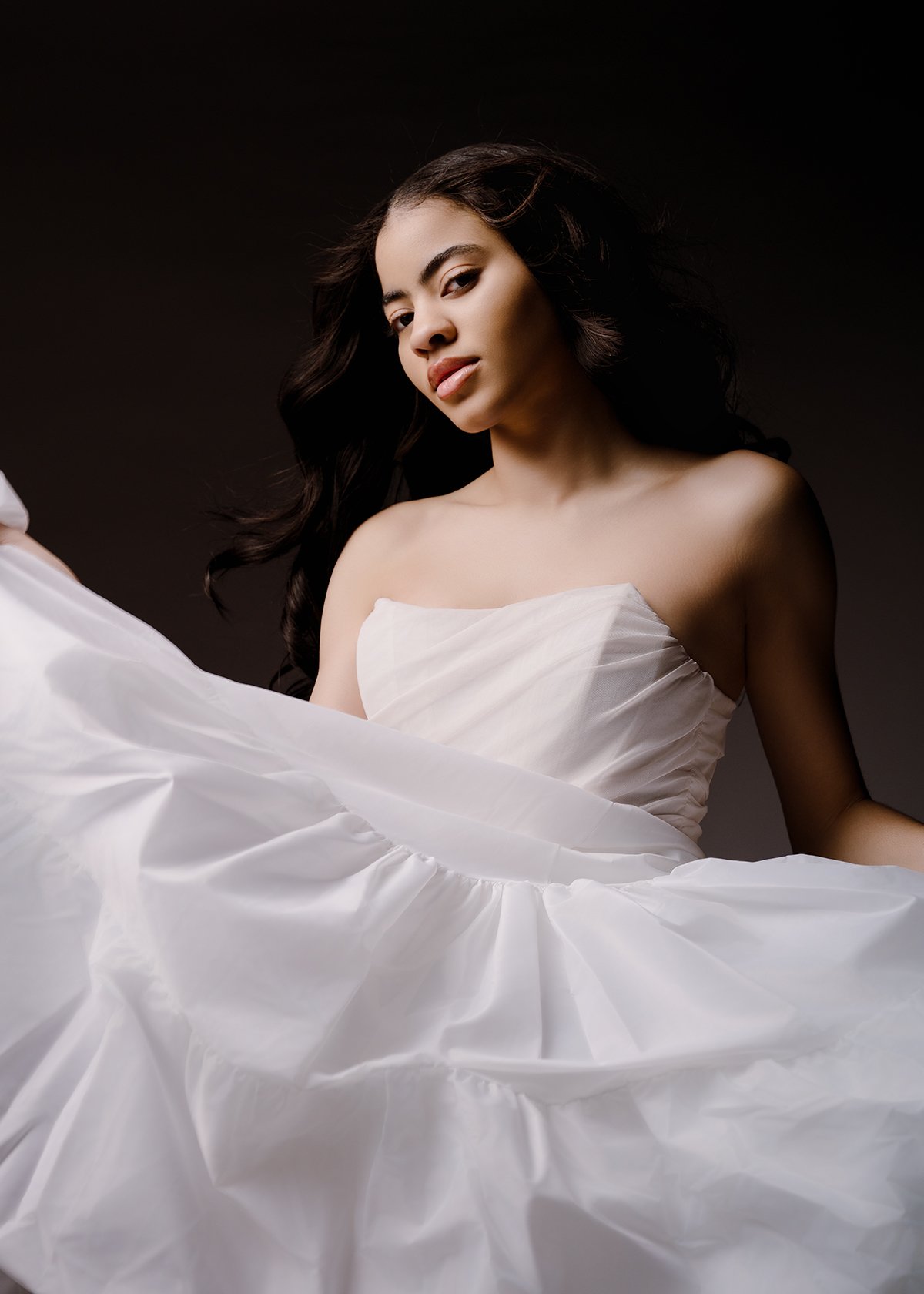 A young woman with long dark hair, wearing a strapless white dress, looks confidently at the camera against a dark background.