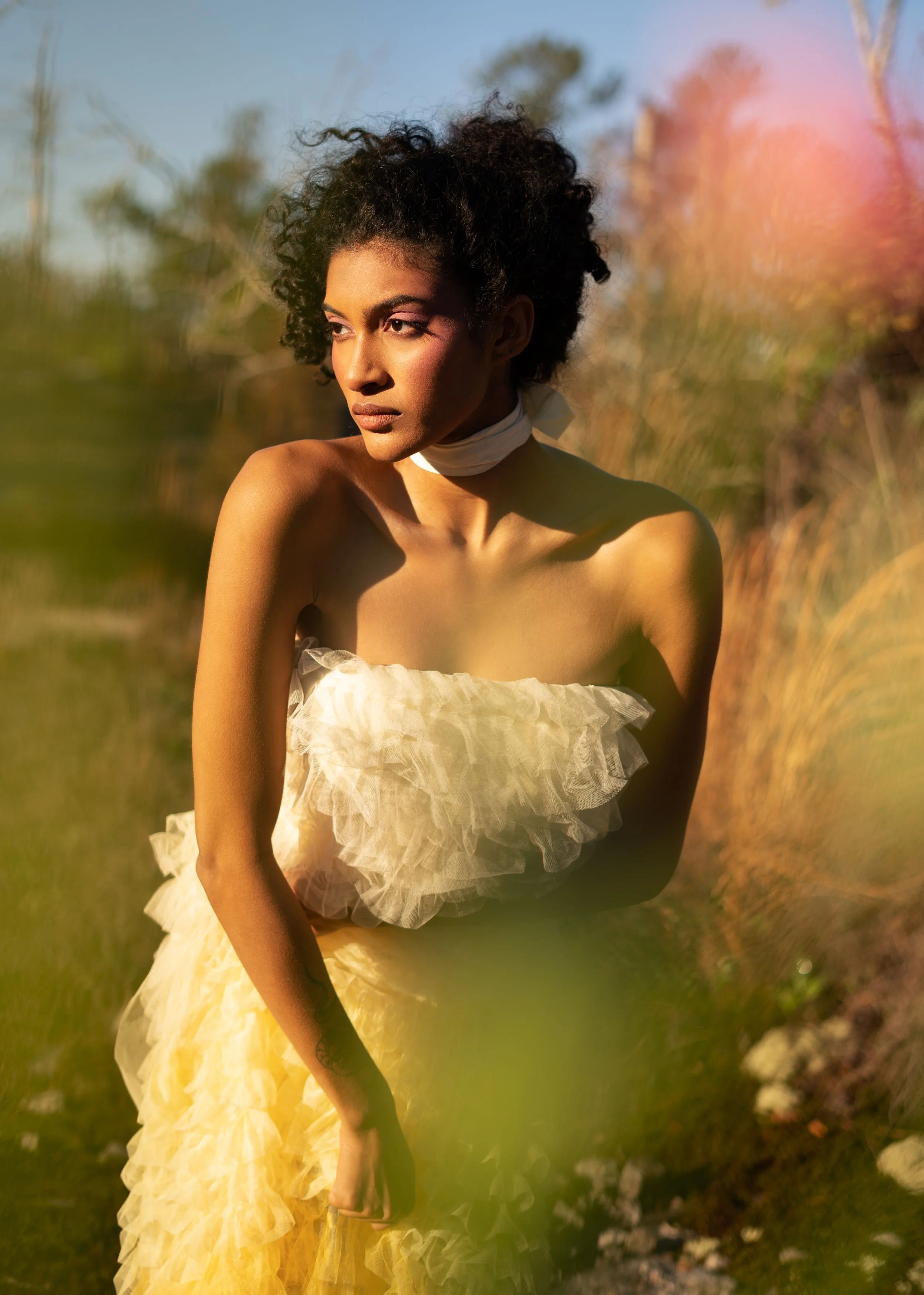 A woman with dark curly hair standing outdoors during golden hour, wearing a strapless cream-colored ruffled dress and a white neck scarf, with a blurred natural background.