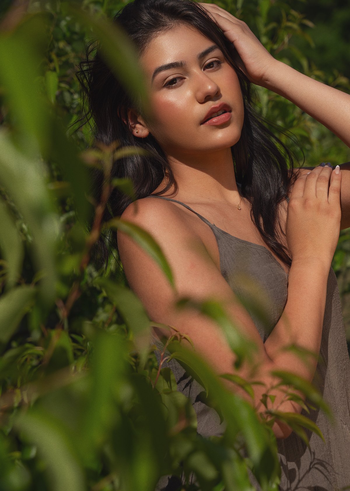 A young woman with long dark hair and light makeup posing outdoors among green foliage, wearing a sleeveless grey top.