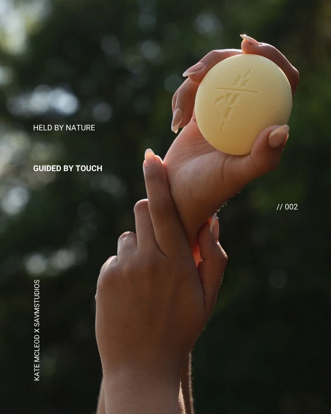 A hand holding a round, yellow soap with the word 'RITUAL' engraved on it, outdoors with blurred green foliage in the background.