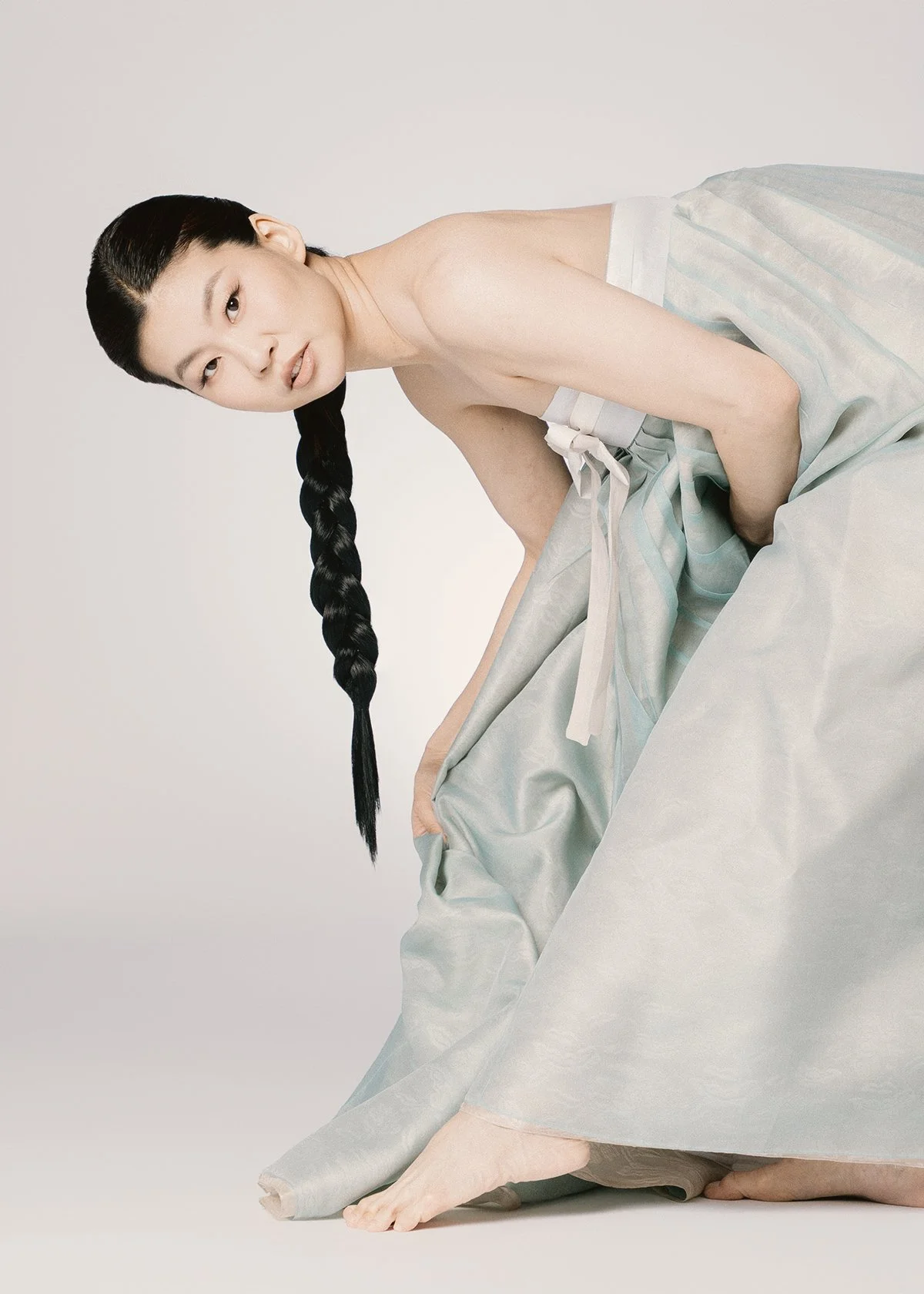 A woman with long black hair in a braid kneeling on the ground. She is topless, wearing a pastel-colored skirt, and gazing at the camera against a plain white background.
