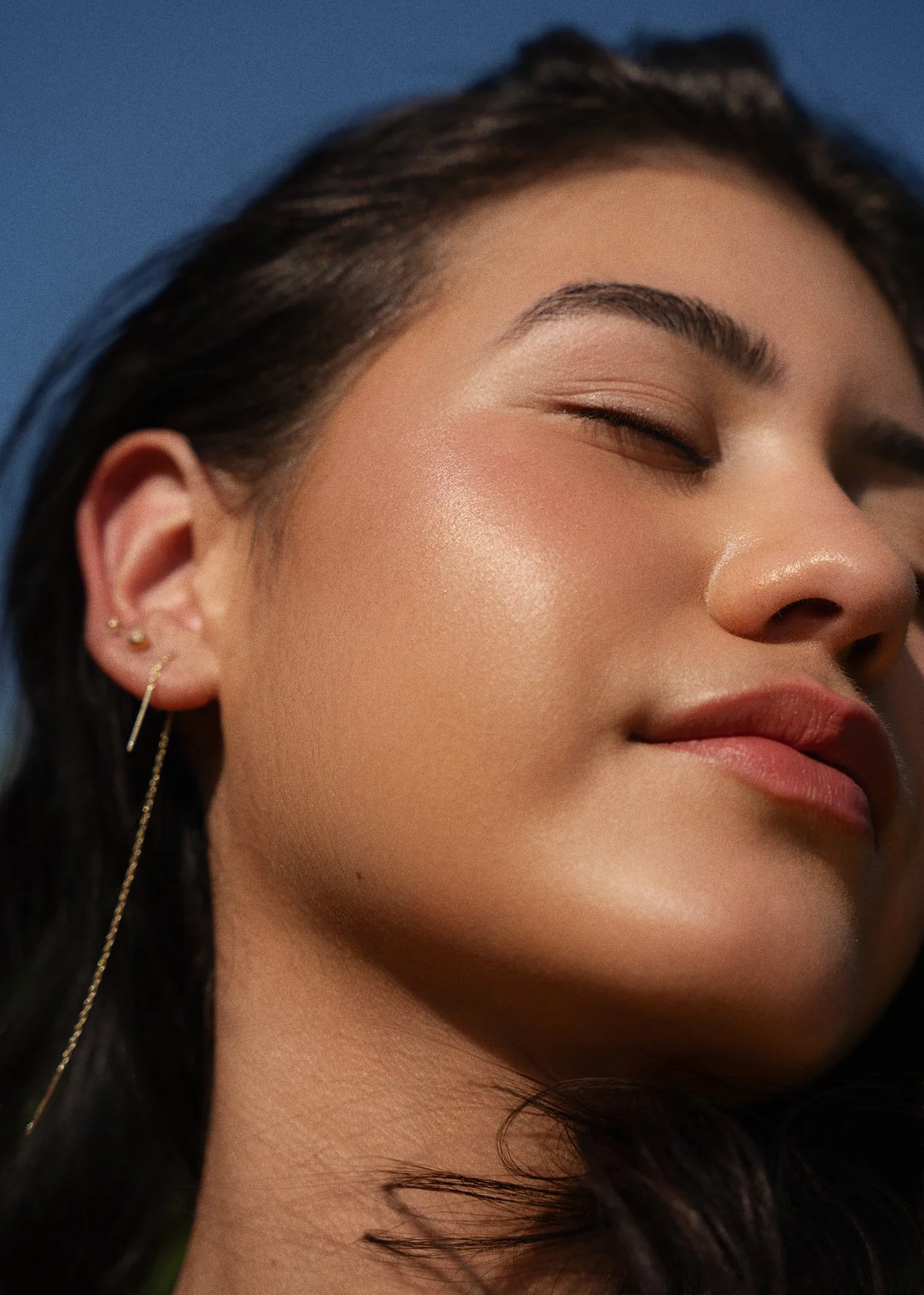 Close-up of a woman with her eyes closed, showing smooth skin, glossy lips, and multiple earrings on her left ear. Background is dark blue.