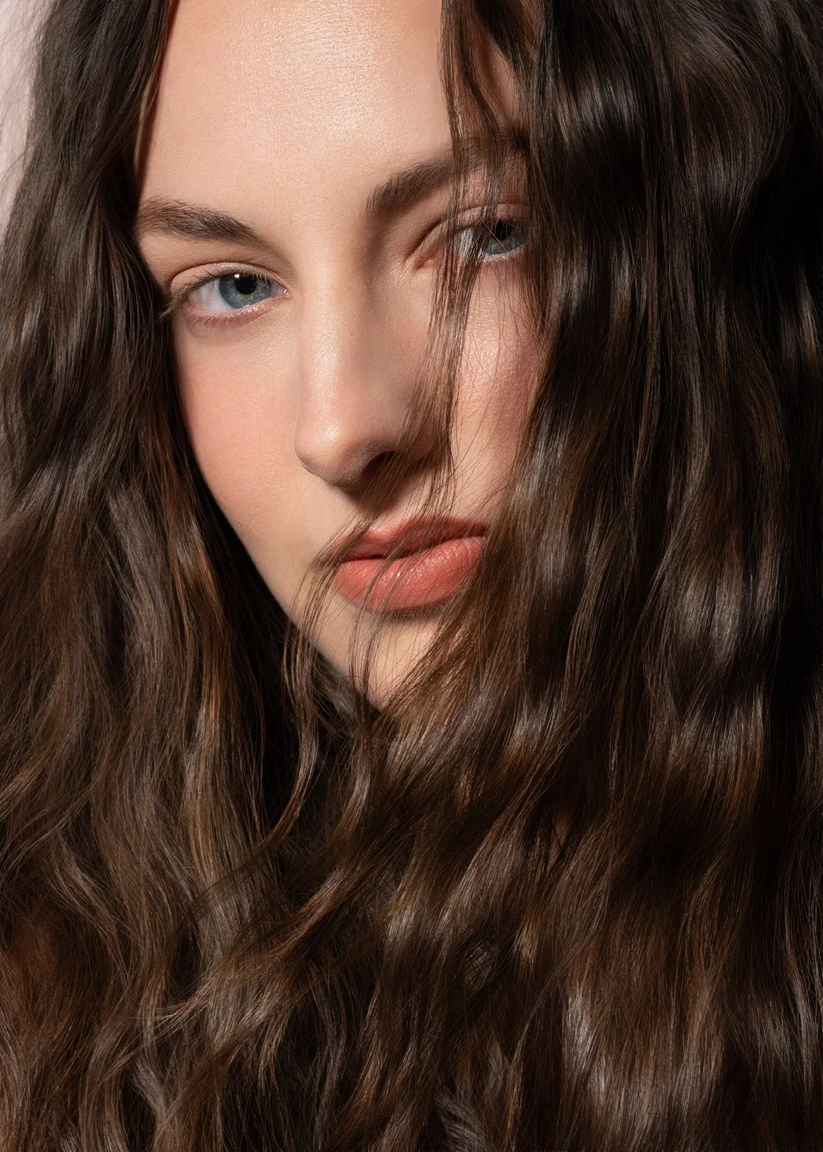 Close-up of a woman with long, wavy brown hair covering part of her face, light skin, blue eyes, and natural makeup.