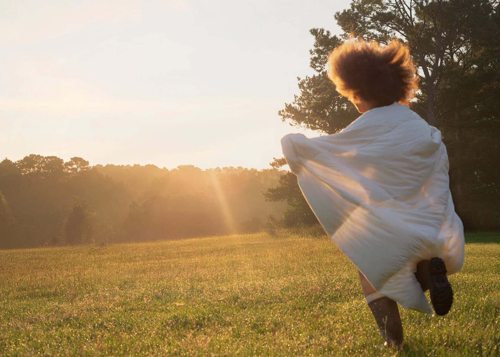 Person with curly hair running across a grassy field during sunset, wearing a light-colored coat or blanket.