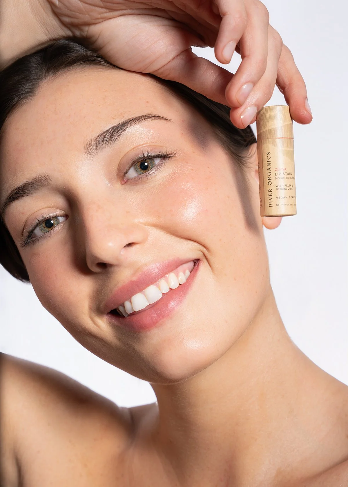 A woman with fair skin and brown hair smiling and holding a lip stain tube near her face