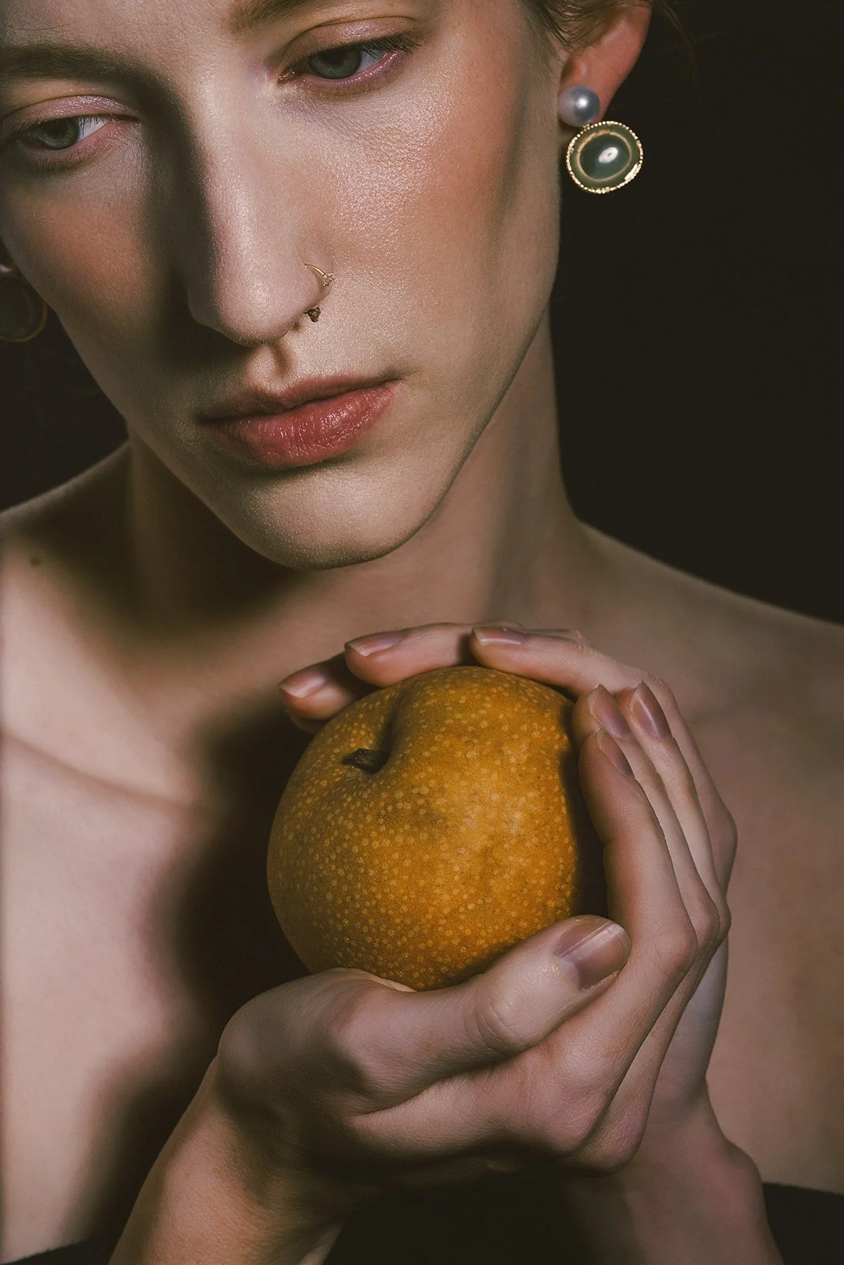 A close-up of a woman with fair skin and blue eyes holding a yellow-orange pear against her chest. She has pearl and decorative earrings, a subtle nose ring, and is looking downward with a calm expression against a dark background.