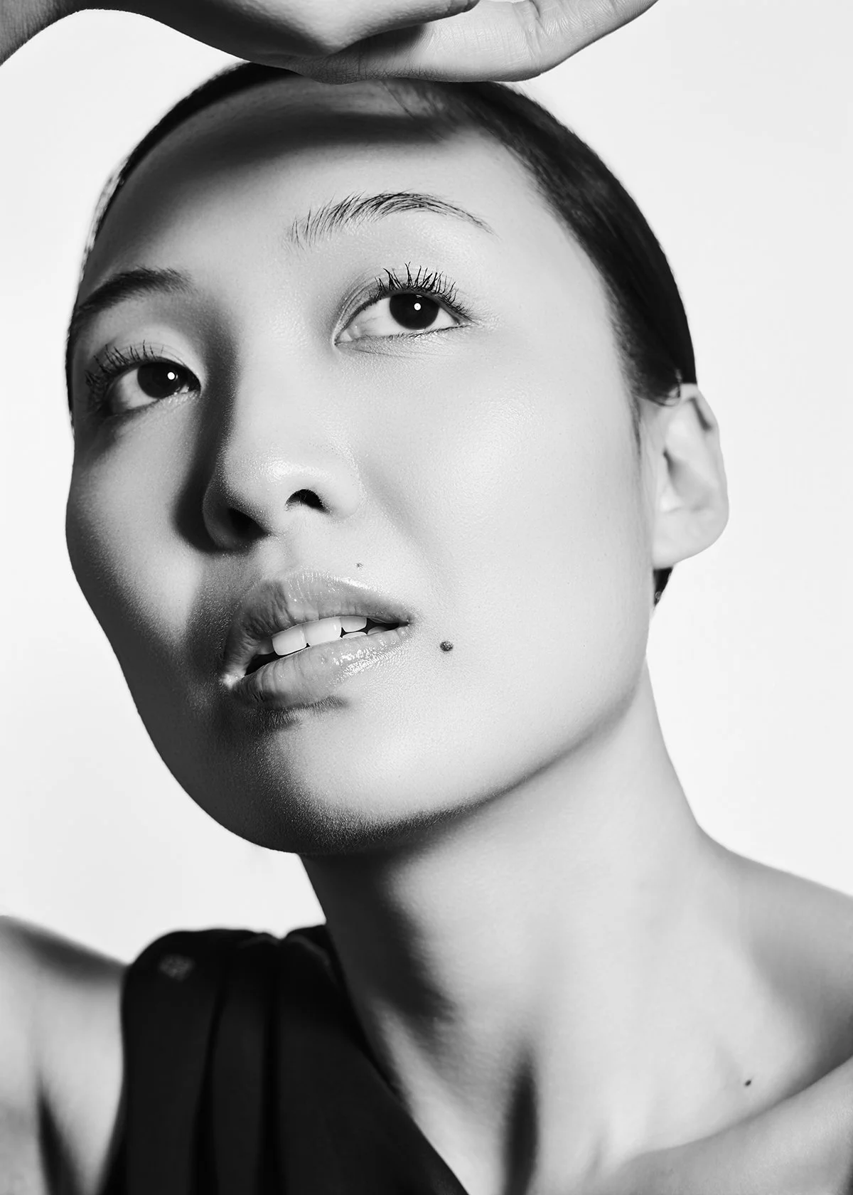 Close-up black and white portrait of a woman with short hair, a contemplative expression, and a beauty mark on her lower lip, holding her hand above her forehead.