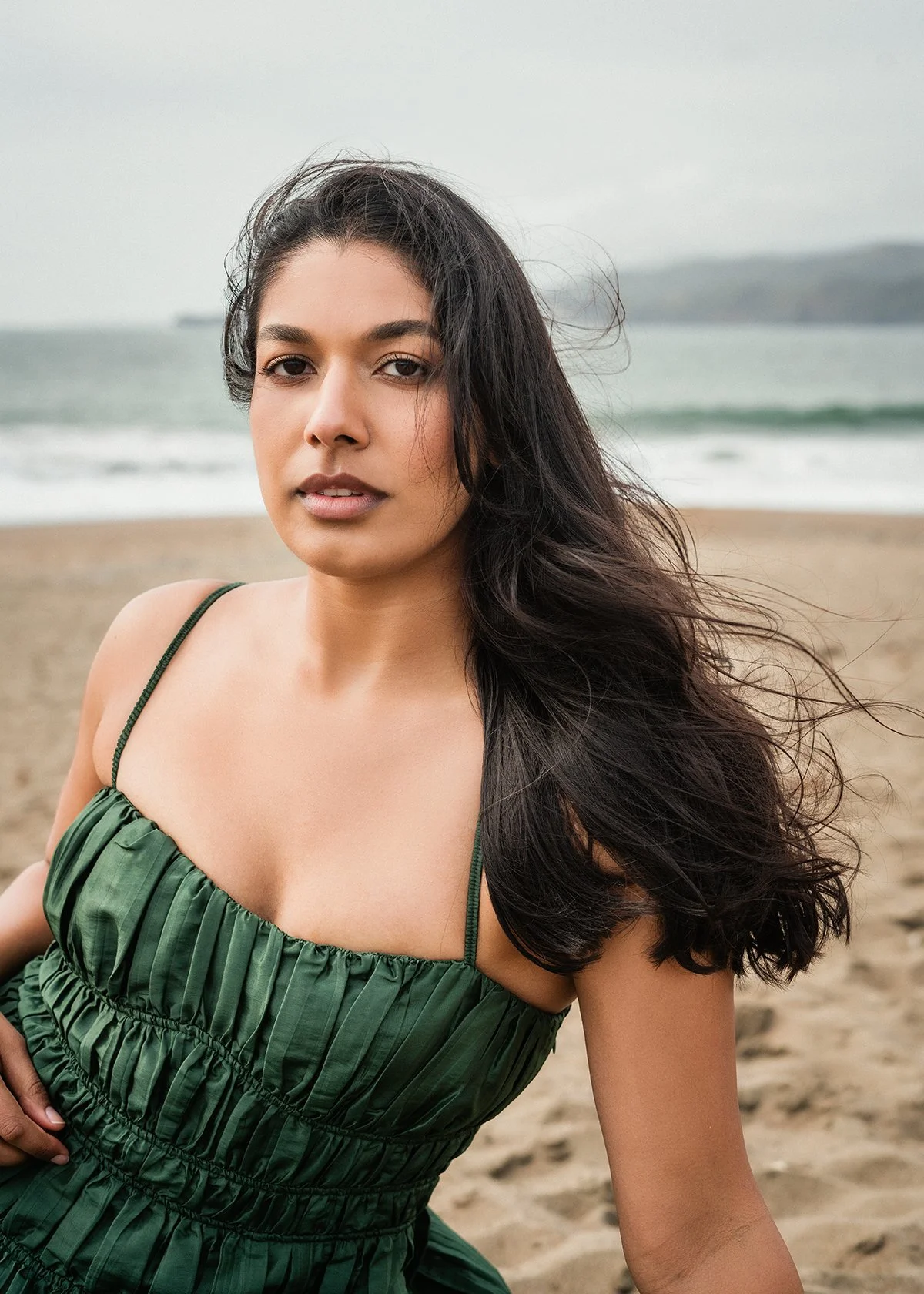 A woman in a green dress sitting on the beach with ocean waves and a cloudy sky in the background.