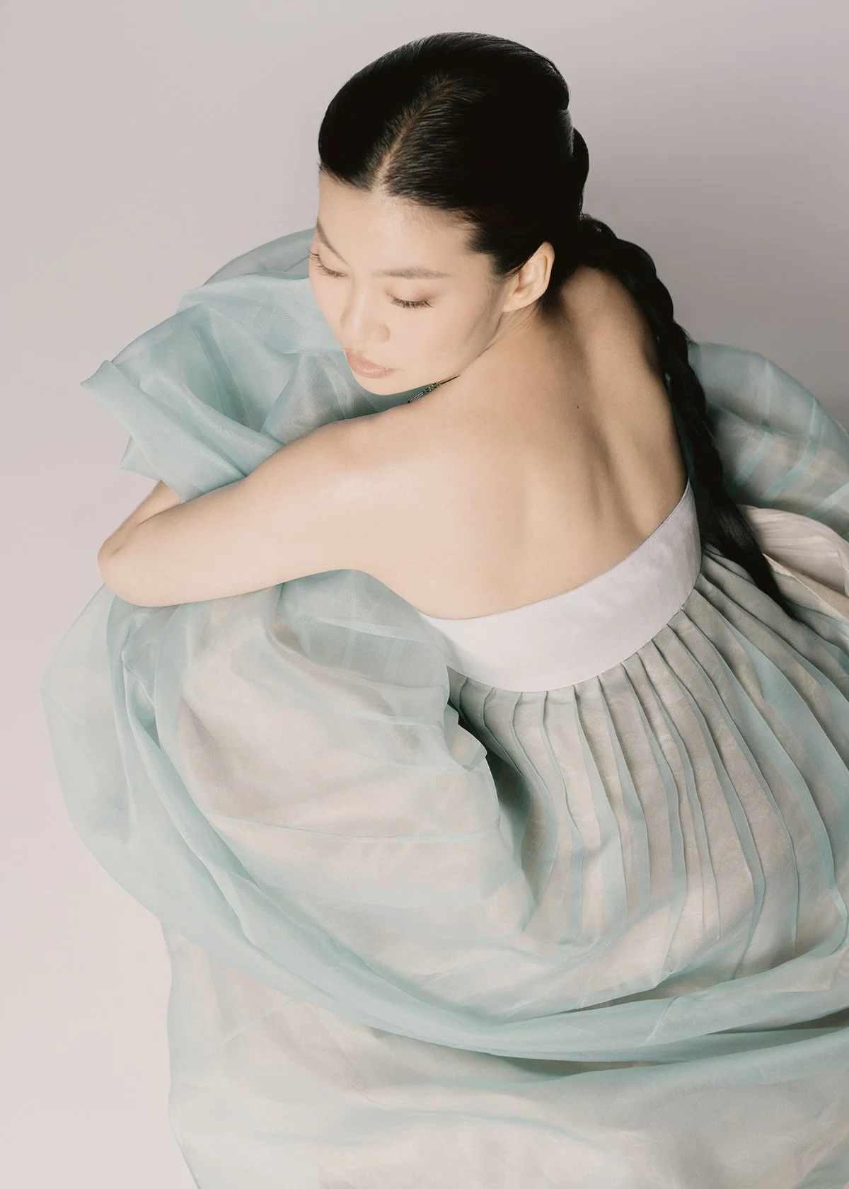 A woman with dark hair styled in a braid, wearing a strapless dress with light-colored fabric, sitting with her arms crossed over her knees, looking down with a serene expression.