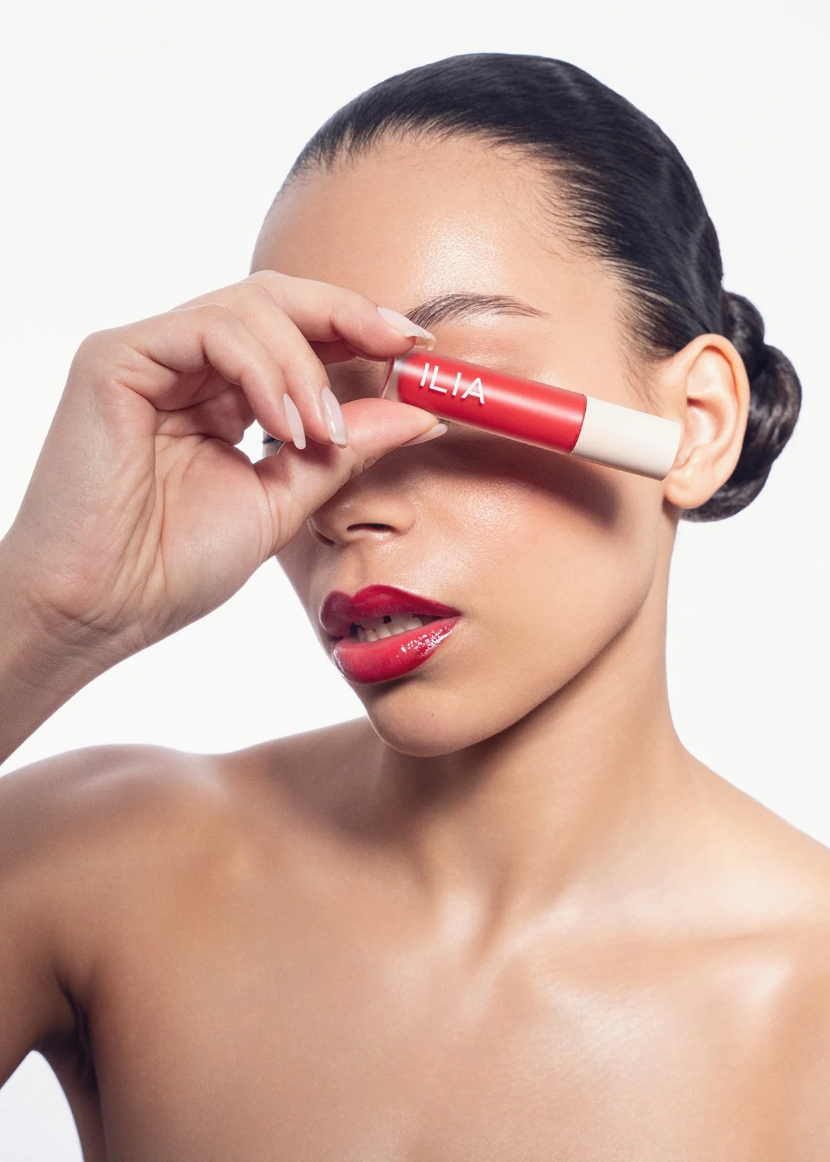 A woman with dark hair styled in a low bun is holding a lip balm called ILIA in front of her face, covering her eyes, against a white background.