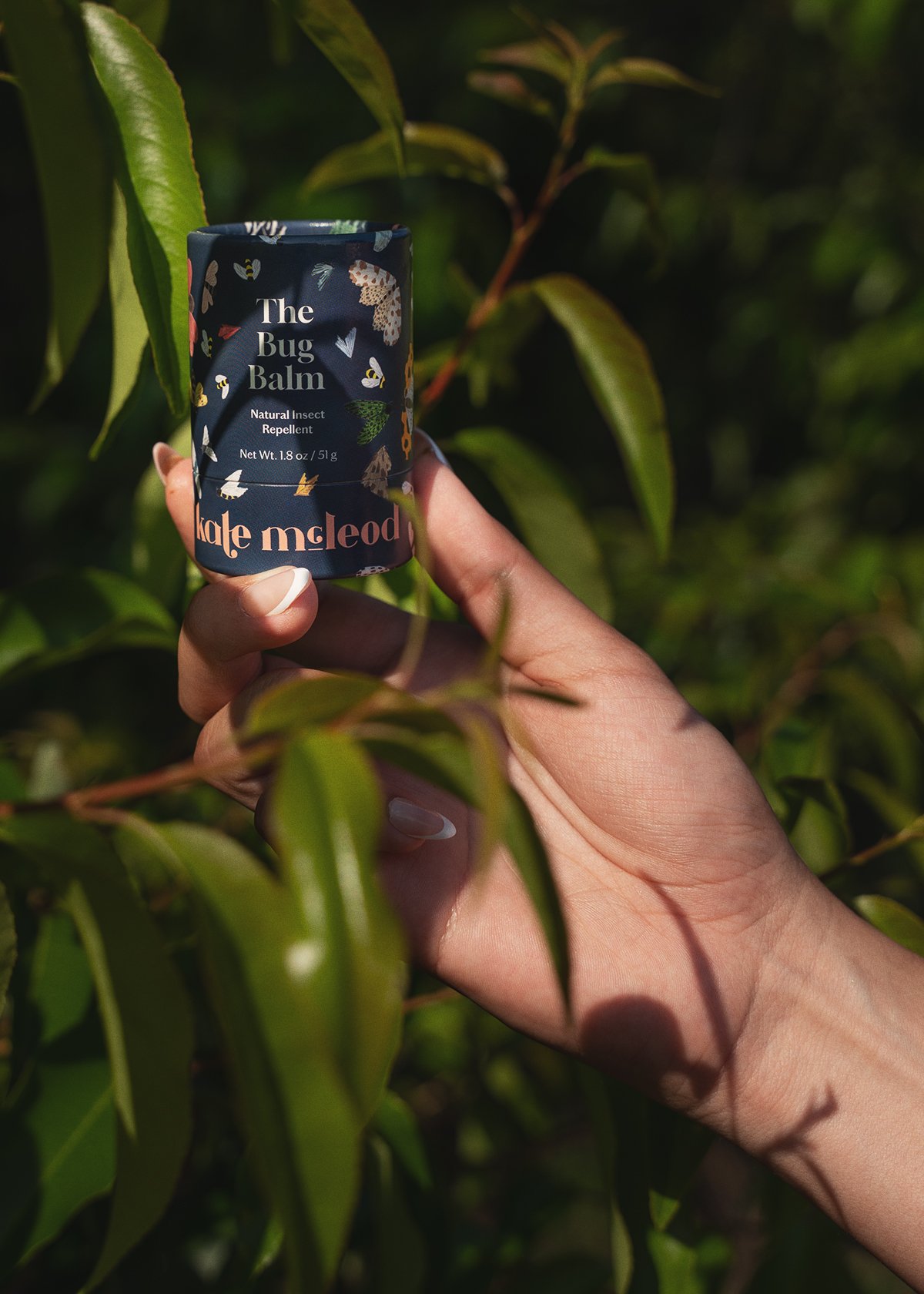 A hand holding a small container labeled "The Bug Balm" among green leaves on a plant.