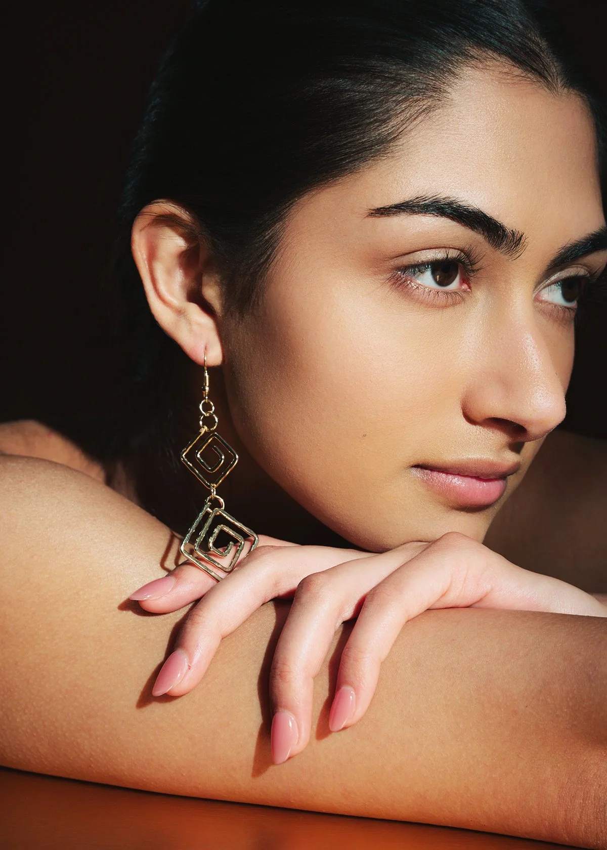 Close-up of a woman with dark hair, wearing large geometric gold earrings, resting her chin on her hand, with smooth skin and natural makeup, against a dark background.