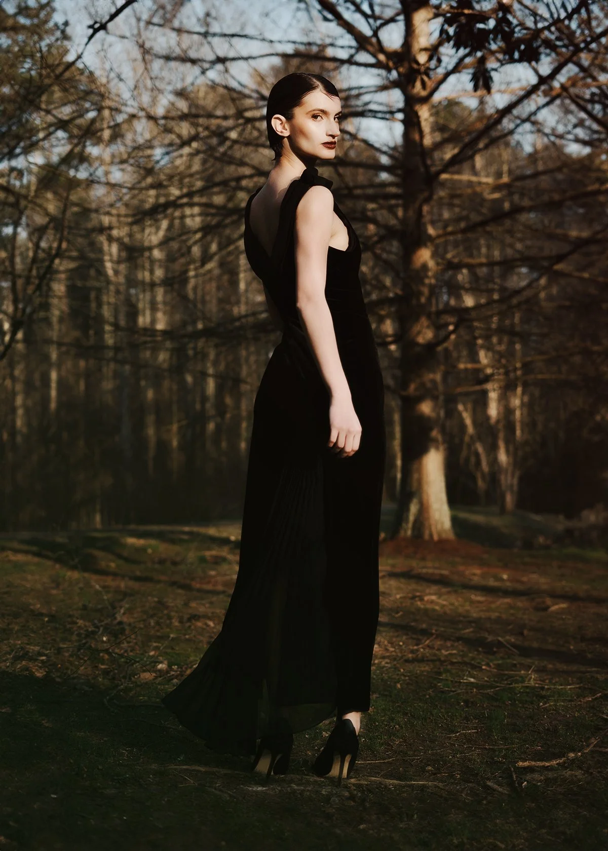 A woman in a black dress and high heels standing outdoors in a forest during sunset.