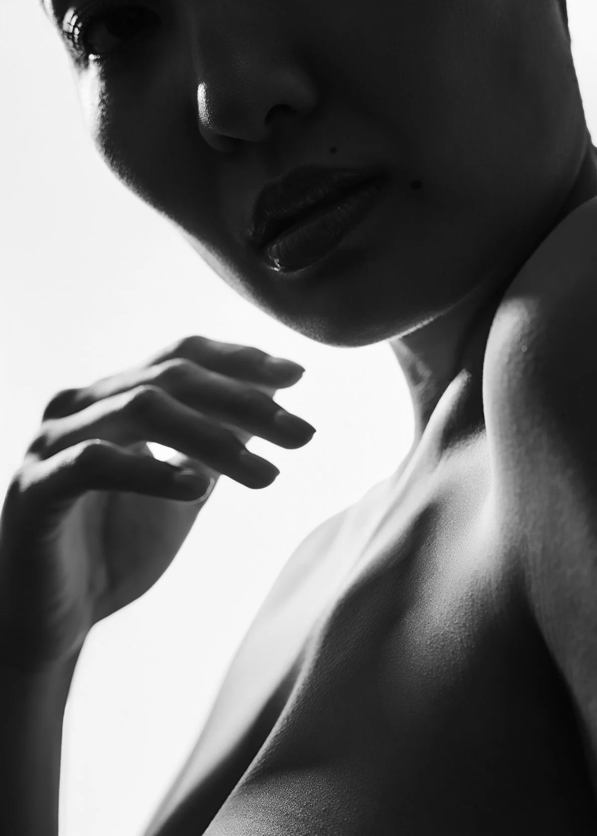 Close-up black and white photo of a woman's face and shoulder, with her hand near her chin, high contrast lighting highlighting facial features and skin texture.