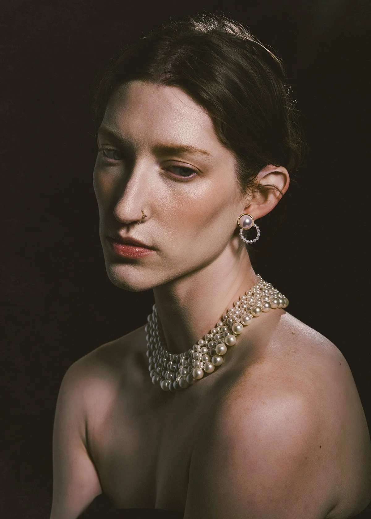 A woman with short dark hair and light skin tone wearing pearl jewelry, including a multi-strand pearl necklace, pearl earrings, and a nose ring, posing against a dark background.