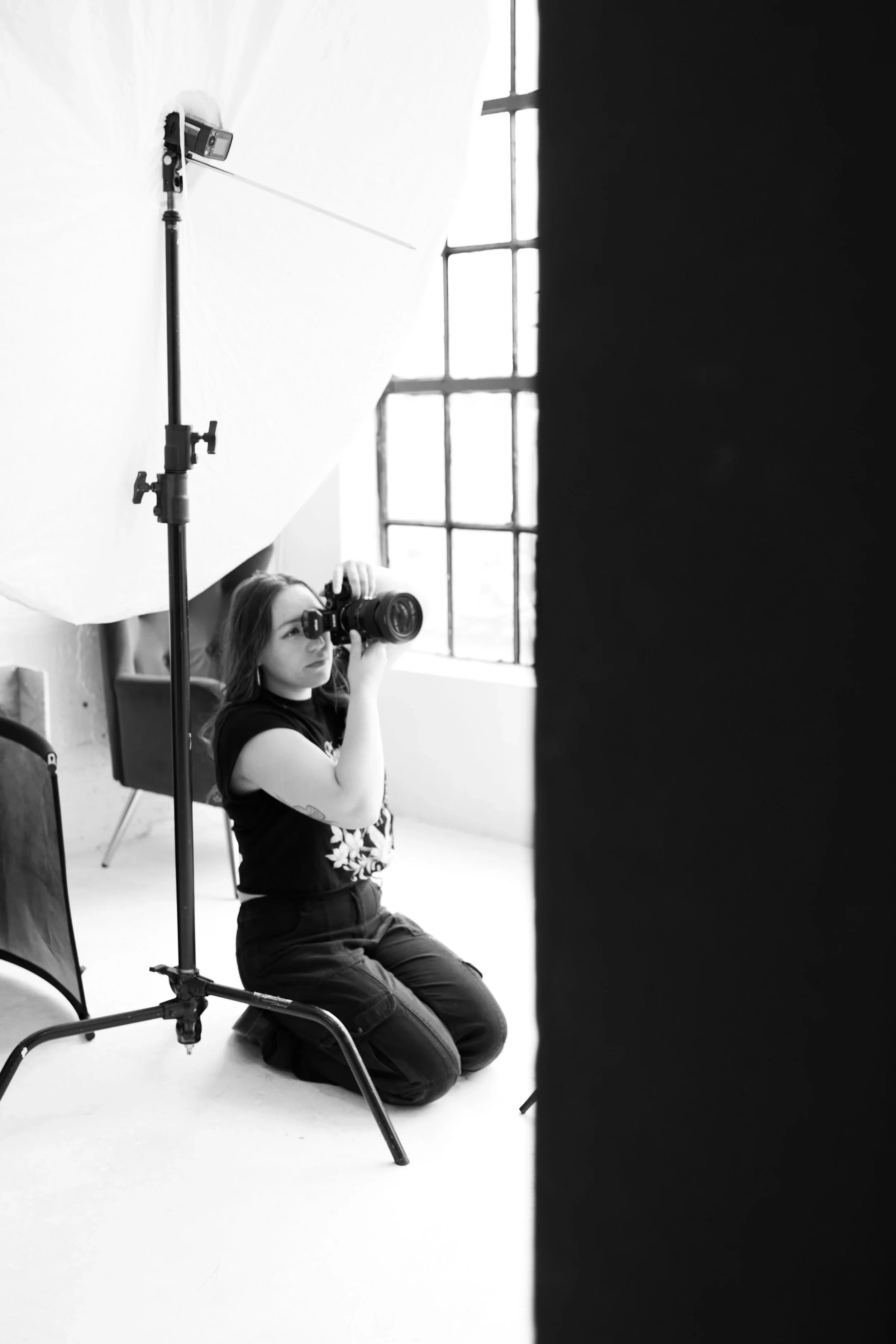 A woman kneeling on the floor in a photography studio taking a photo with a DSLR camera, with studio lighting equipment and a large softbox in the background, and a window to the right.