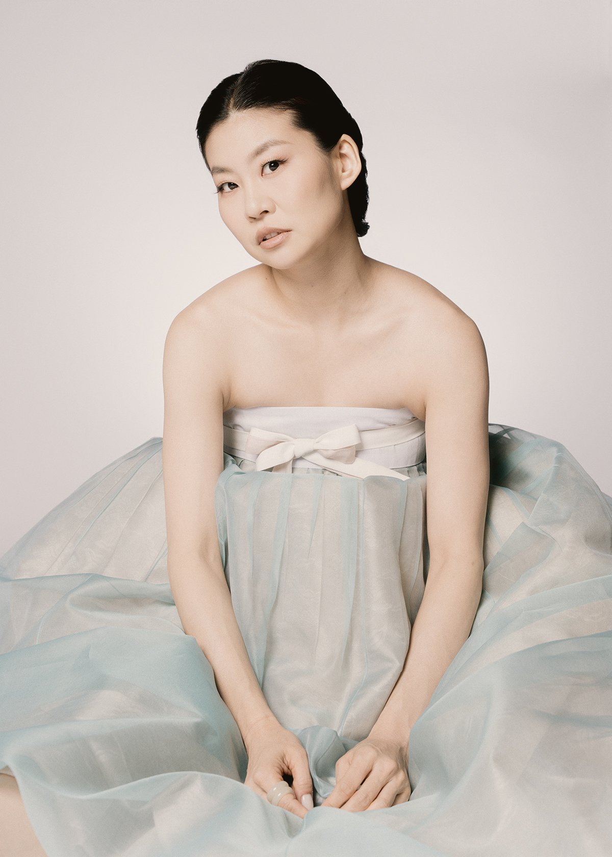 A woman with short black hair and light skin, sitting with her arms resting on her knees, wearing a strapless dress with a ribbon bow on the bodice and a flowing skirt with soft pastel colors, against a plain light background.