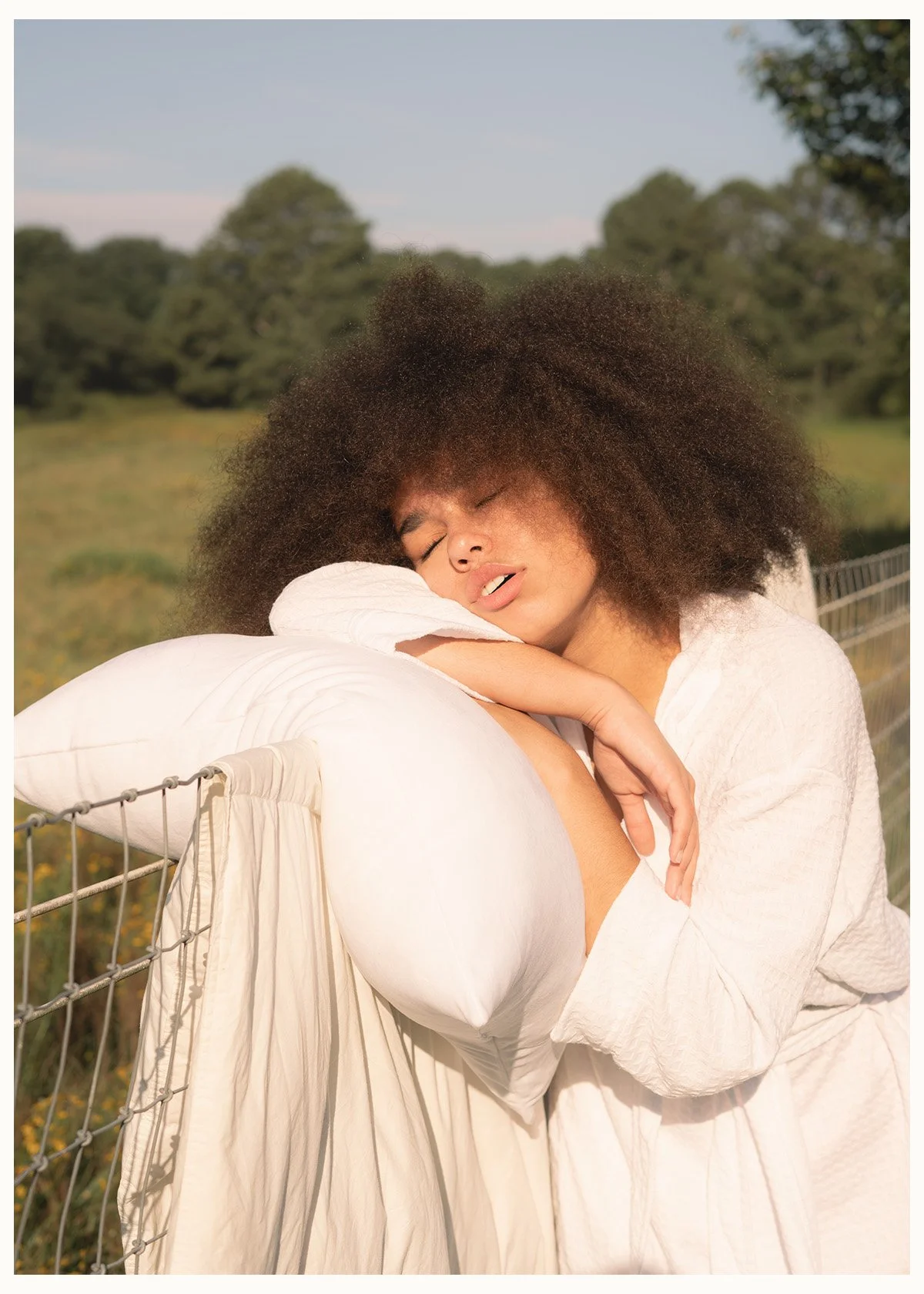A woman with curly hair, wearing a white dress, leaning on a white pillow and gazing with closed eyes outdoors near a fence, with a green field and trees in the background on a sunny day.
