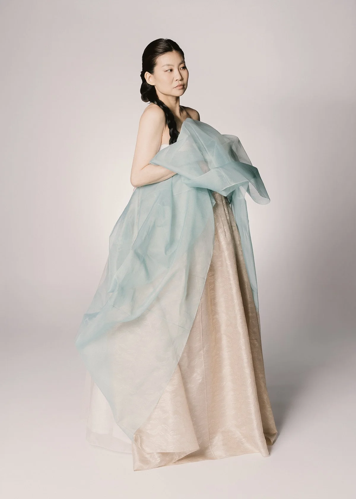 A woman wearing an elegant, historic gown with a beige full skirt and a sheer, light blue shawl draped over her shoulders, posing against a plain light background.