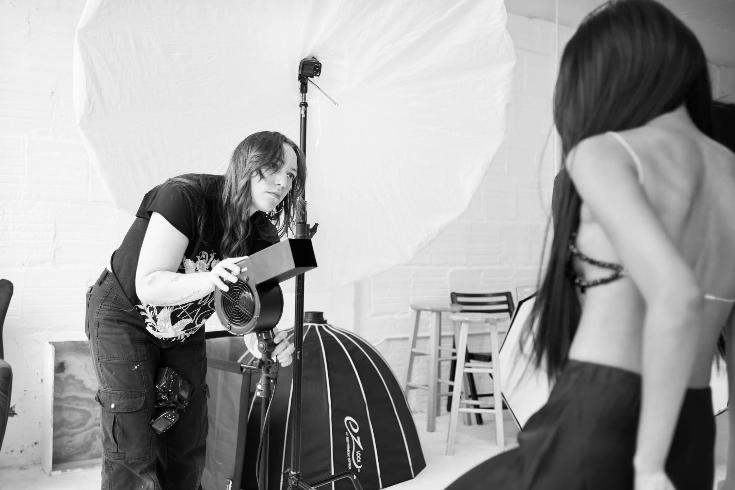 A woman with long hair and a black t-shirt is adjusting a professional photography light with a reflector in a studio setting. Another person with long hair is partially visible, facing her, in the foreground.
