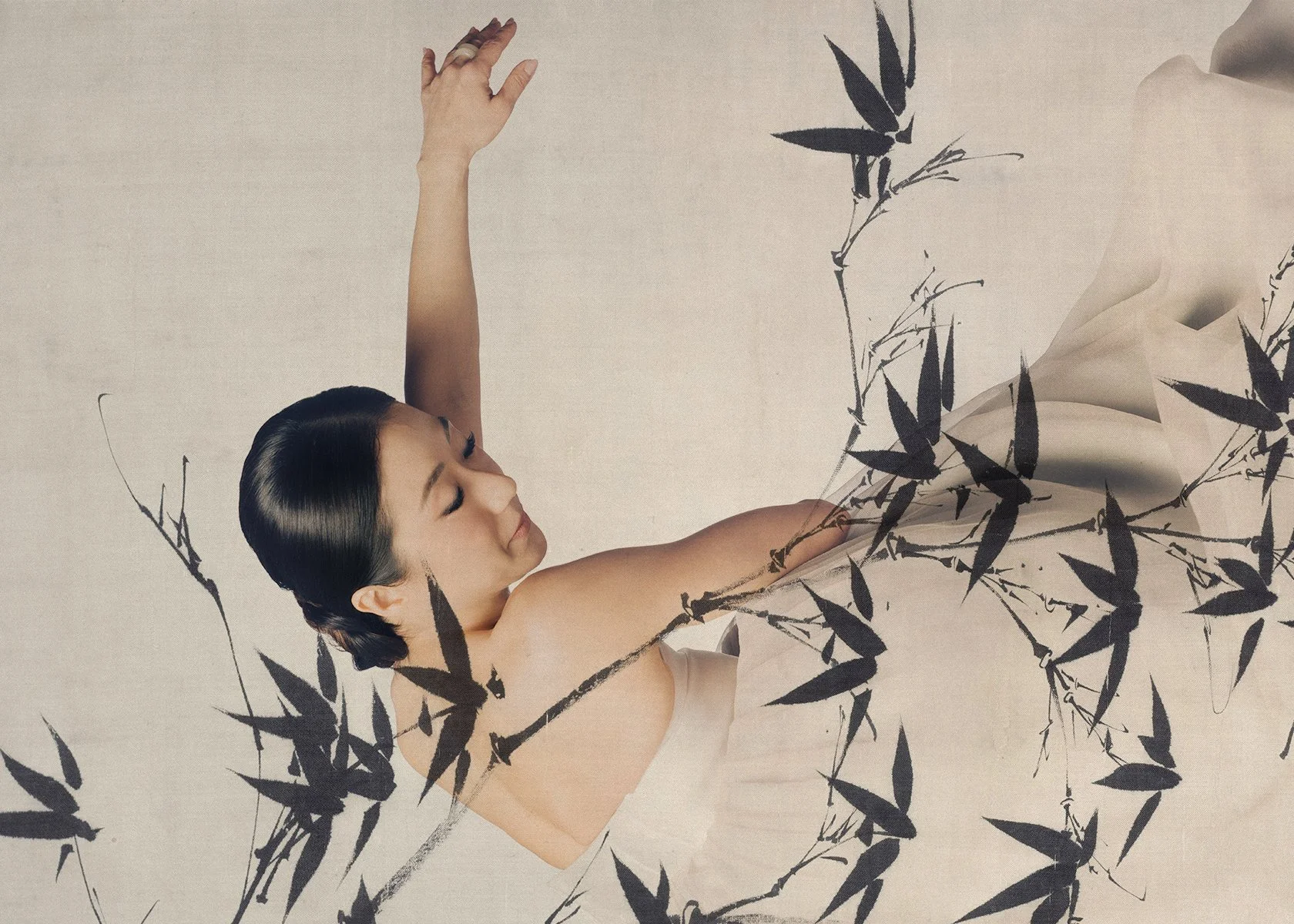 A woman with black hair in a bun, lying on her back with eyes closed, surrounded by black ink brushstroke leaves on a beige background.