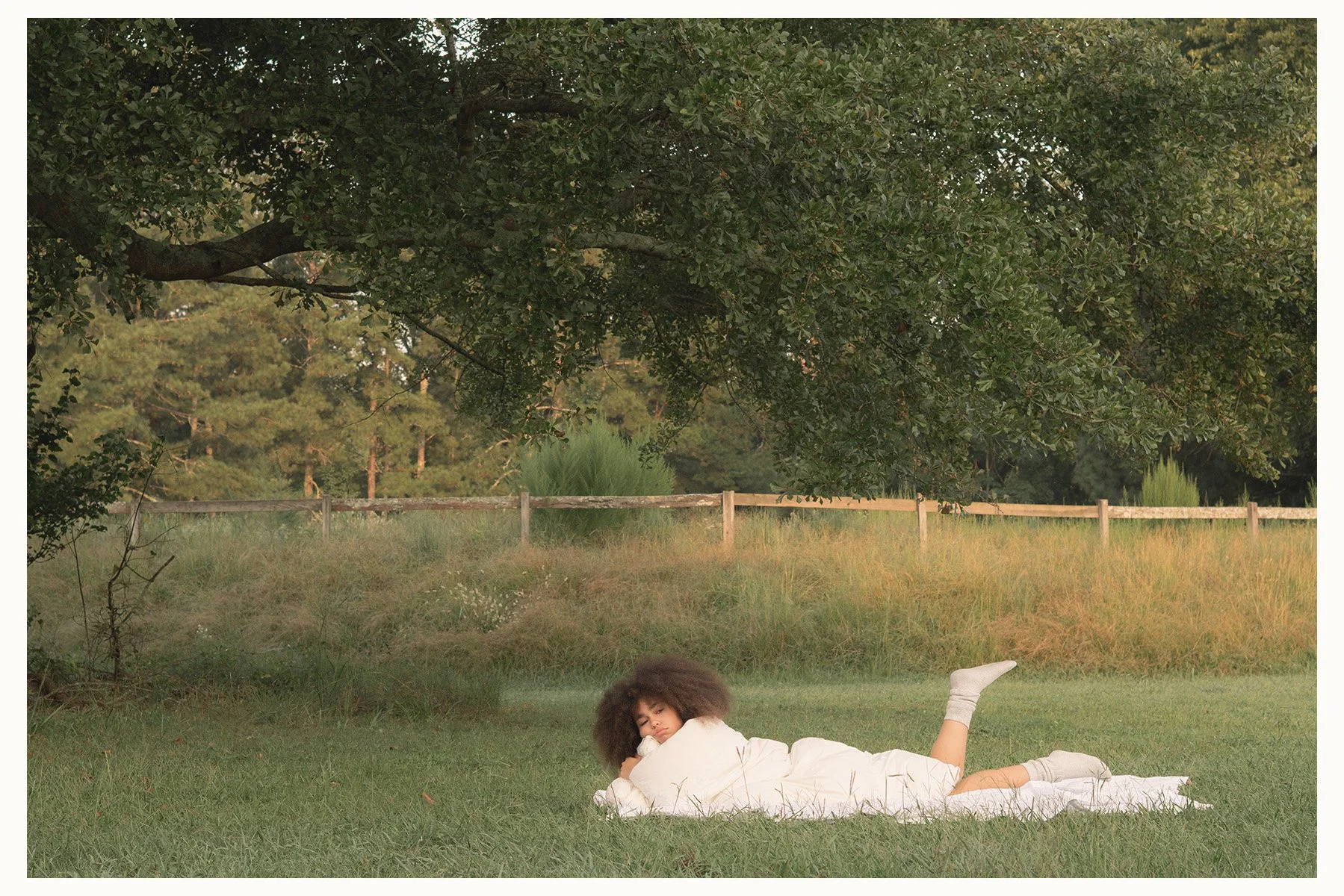 A woman with curly hair lying on her side on a grassy field, under a large tree, resting her head on her arm, wearing a white dress and socks, with a wooden fence and trees in the background.