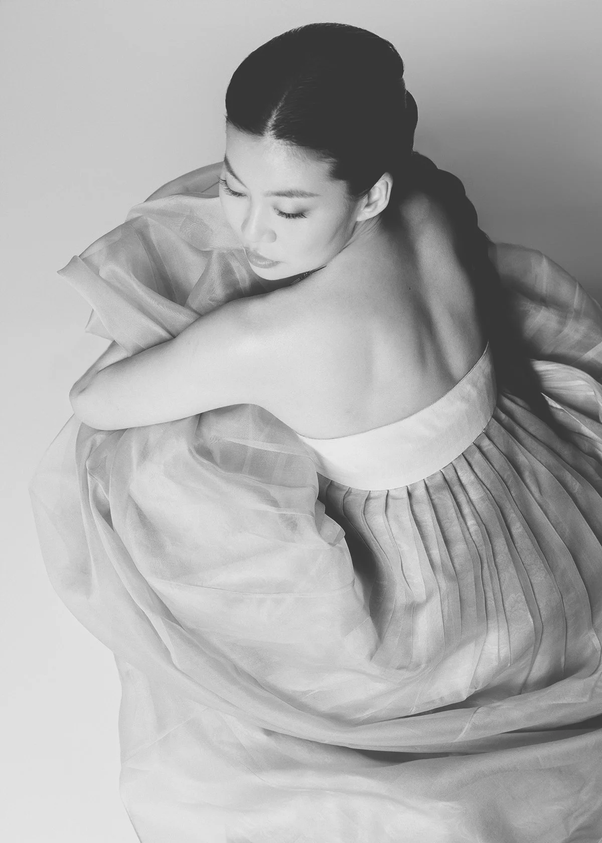 Black and white photo of a woman with a braid, wearing a strapless pleated dress, hugging herself with closed eyes.