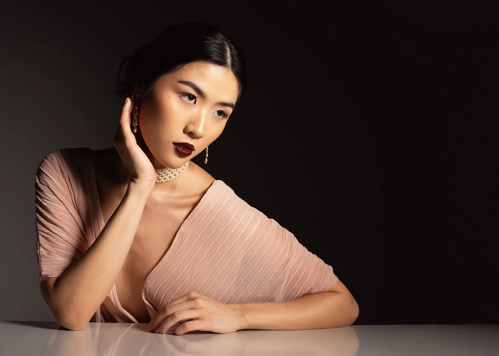 A young woman with dark hair styled in an elegant updo, wearing dark lipstick, pearl jewelry, and a pink dress with a deep neckline, posing against a black background.