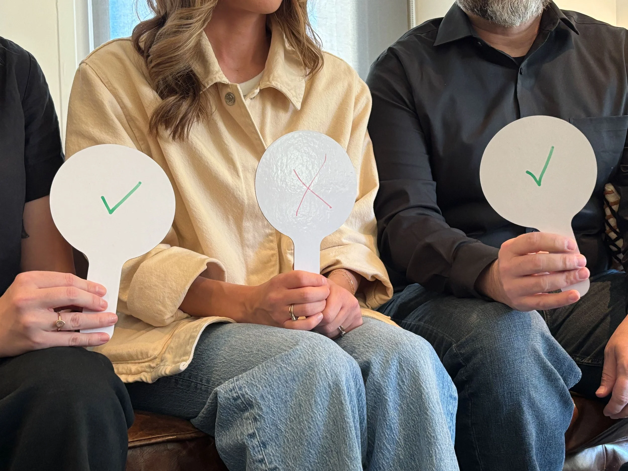 Three people sitting on a couch, each holding a paper speech bubble with a check mark or an X. The person in the middle has no check mark or X. The person on the left has a check mark, the person in the middle has an X, and the person on the right has a check mark.