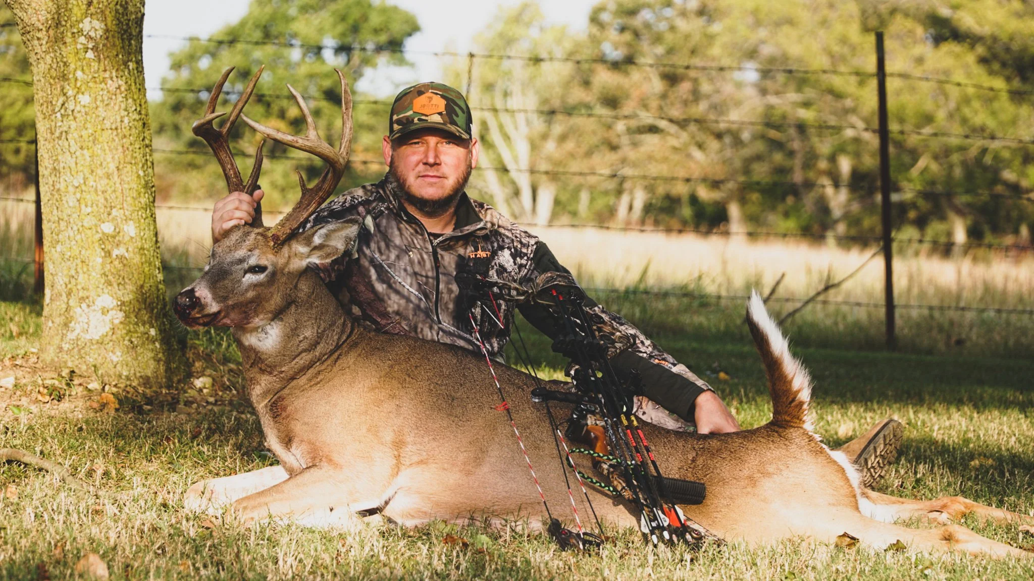 A man in camouflage clothing sitting on grass, holding a large deer with antlers, next to a tree, in an outdoor setting with a fence and trees in the background.