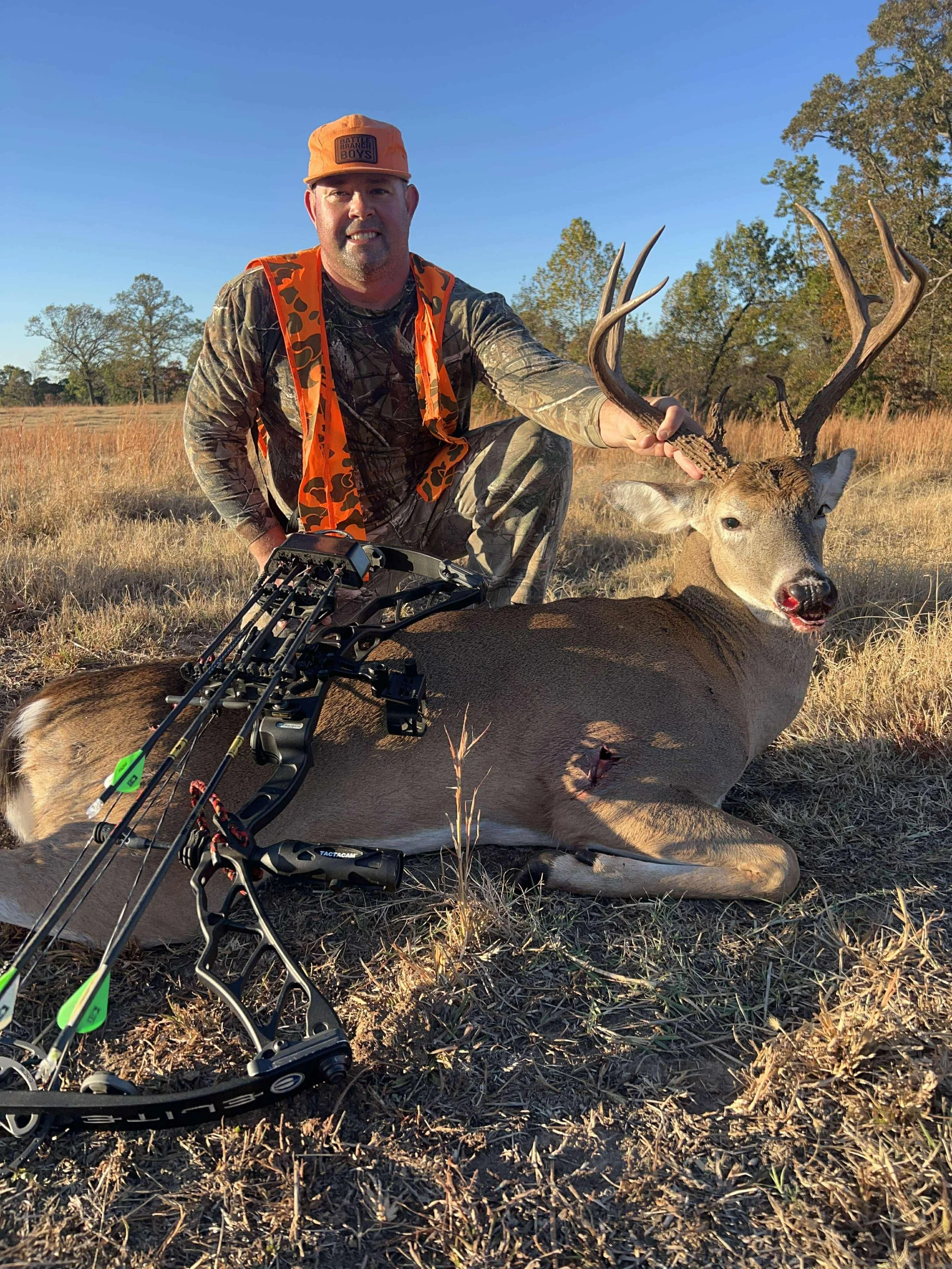 Man in camouflage clothing and orange cap kneels next to a large deer with antlers, holding its antlers, beside a compound bow on the ground in an open field with trees in the background.
