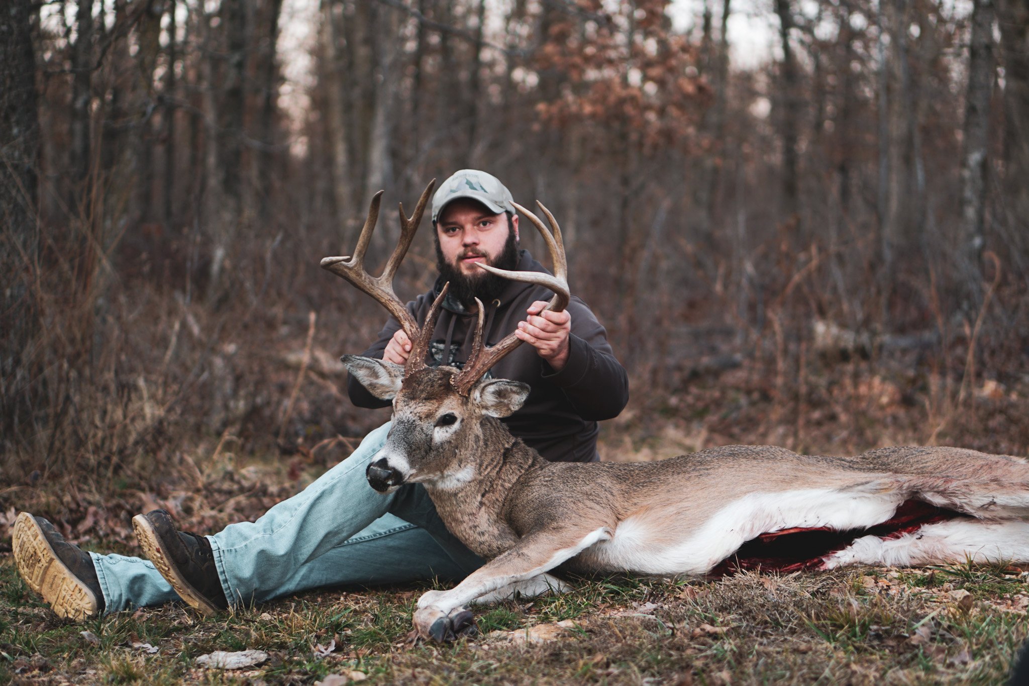A man with a beard wearing a camouflage cap and black hoodie sitting on the ground in a forest, holding the antlers of a recently hunted deer with large antlers, lying on its side with visible wound.