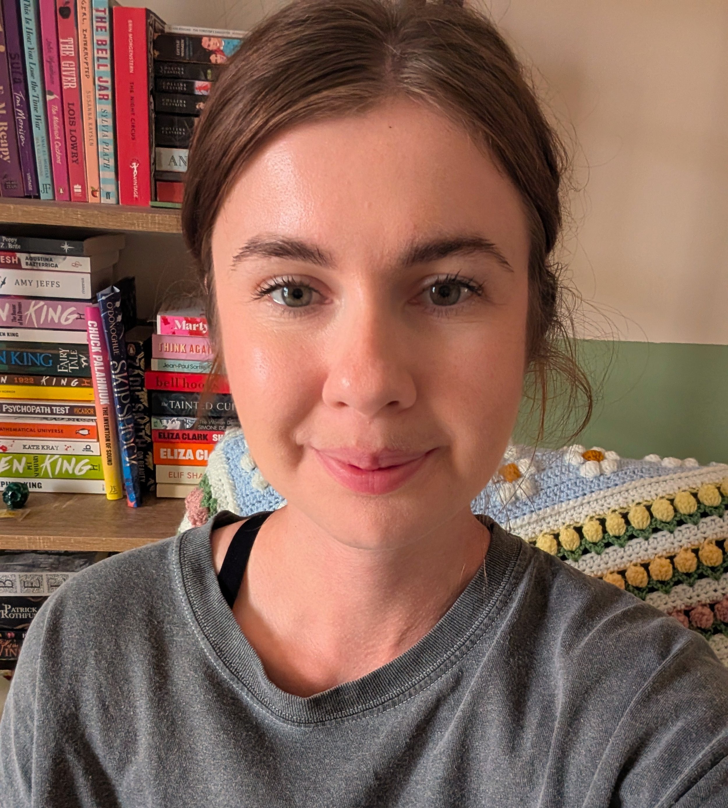 A young woman with light brown hair in a loose bun and light skin, smiling slightly, sitting in front of a bookshelf filled with colorful books.