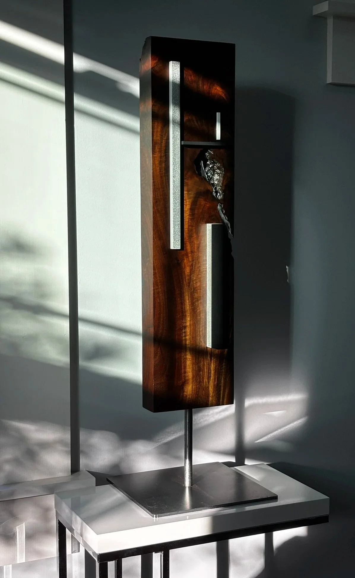 Modern wooden and metal sculpture displayed on a stand with shadows cast on the wall.
