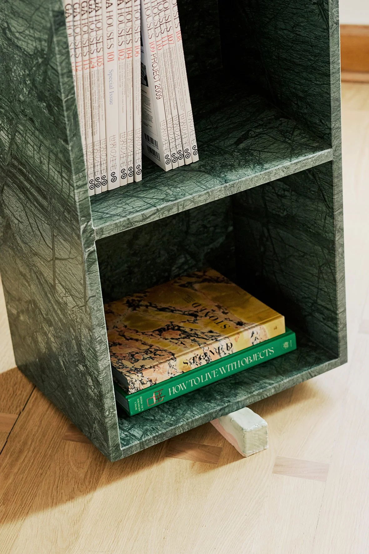 A green marble-patterned bookshelf with two shelves, holding magazines on the top shelf and books on the bottom shelf, placed on a wooden floor.
