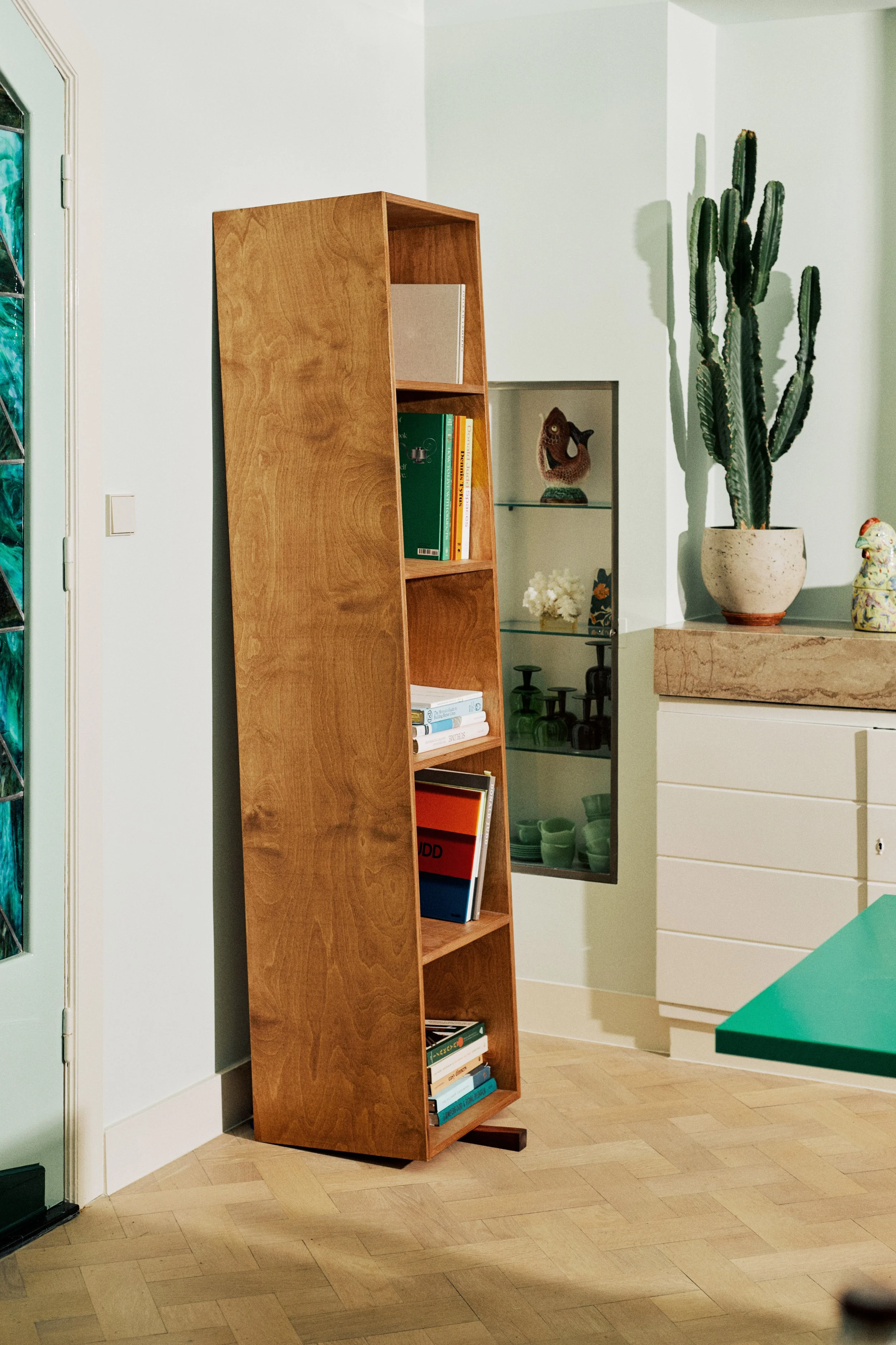 A wooden bookshelf leaning against a wall beside a glass display case filled with decorative objects, with a tall cactus in a pot placed on a surface nearby.