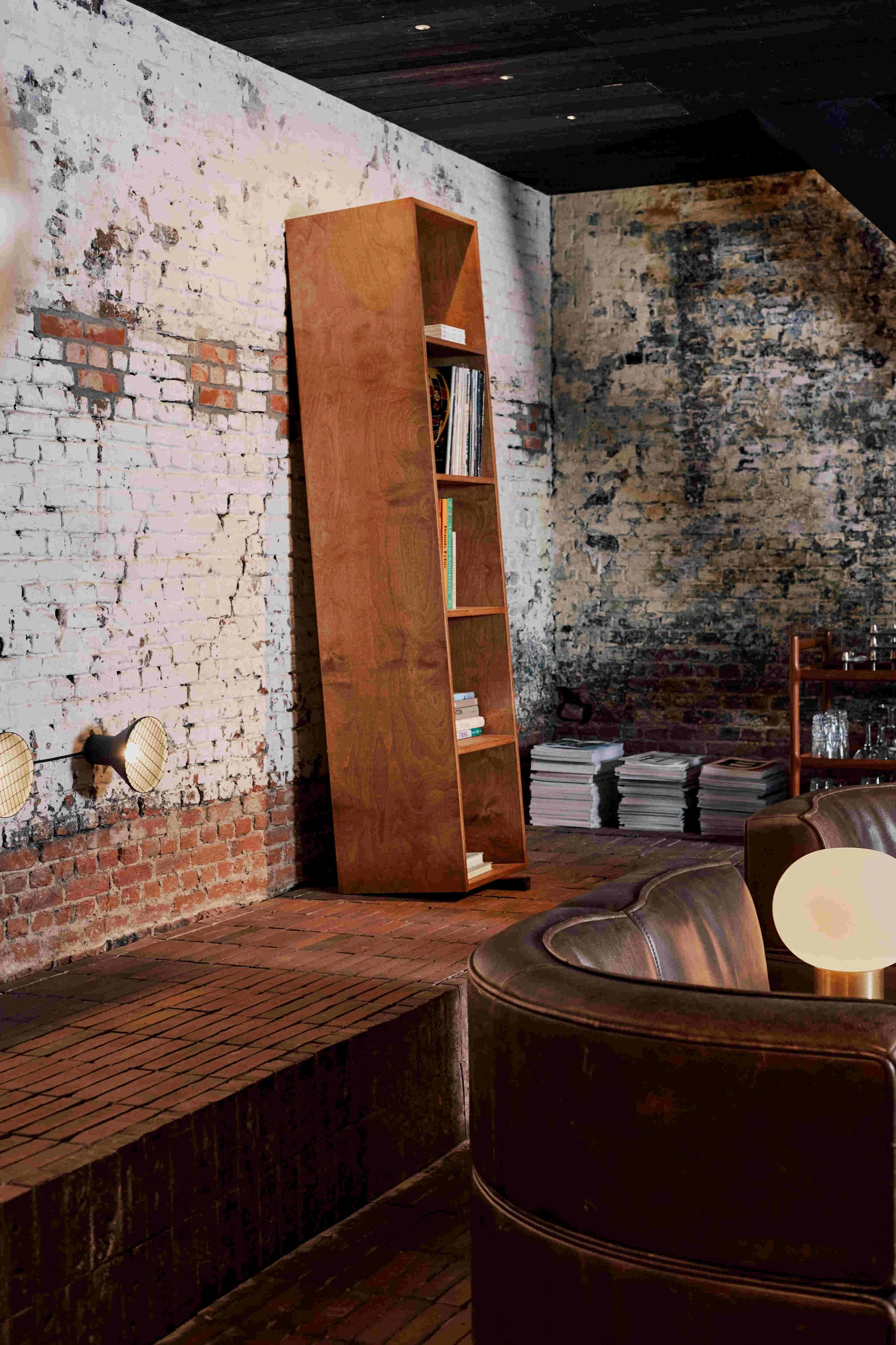Interior of a rustic room with exposed brick wall, wooden bookshelf with magazines, stacks of magazines on the floor, and leather furniture with a small lamp on a table.