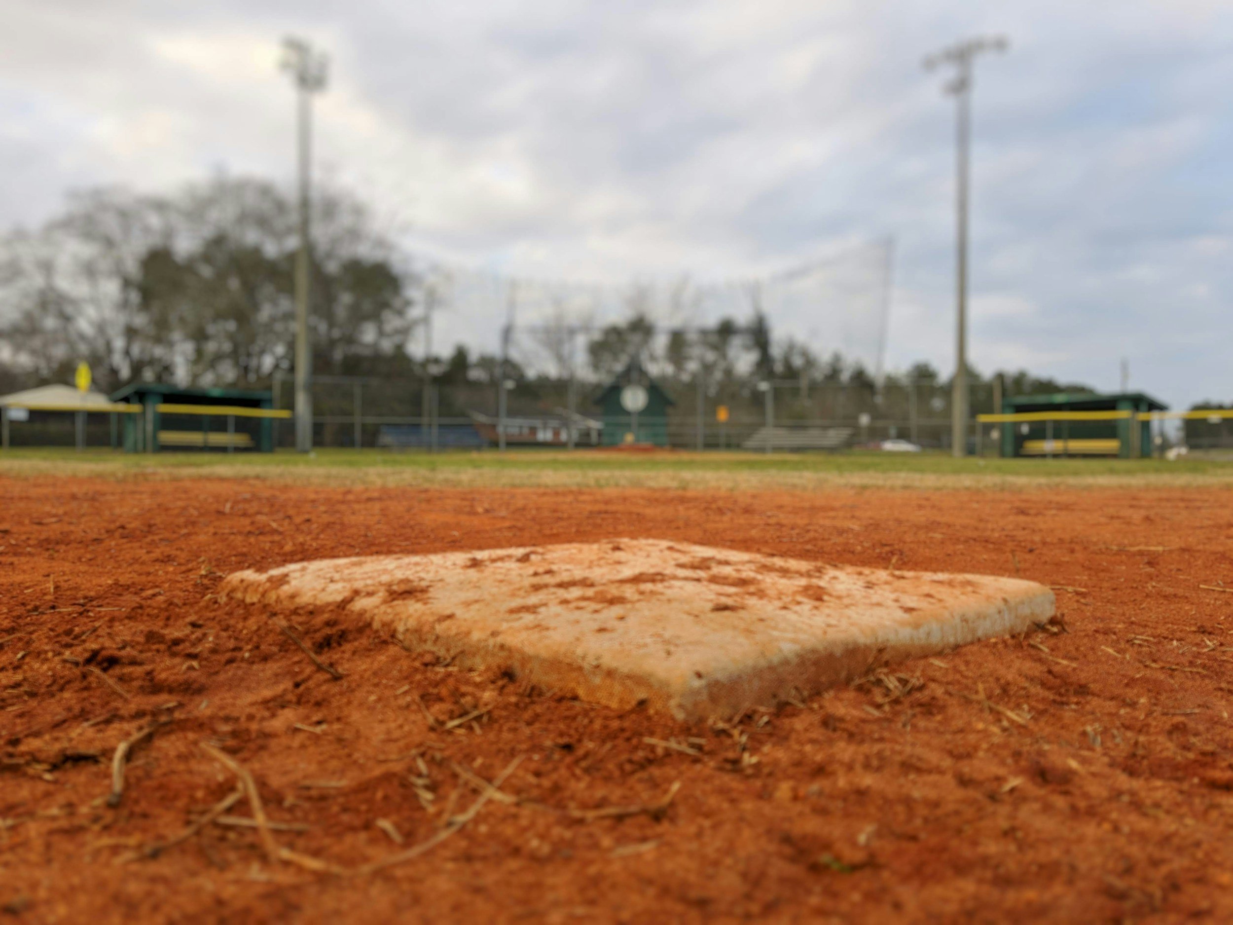 Close-up of a baseball home plate on a dirt field with a blurry baseball field and trees in the background.