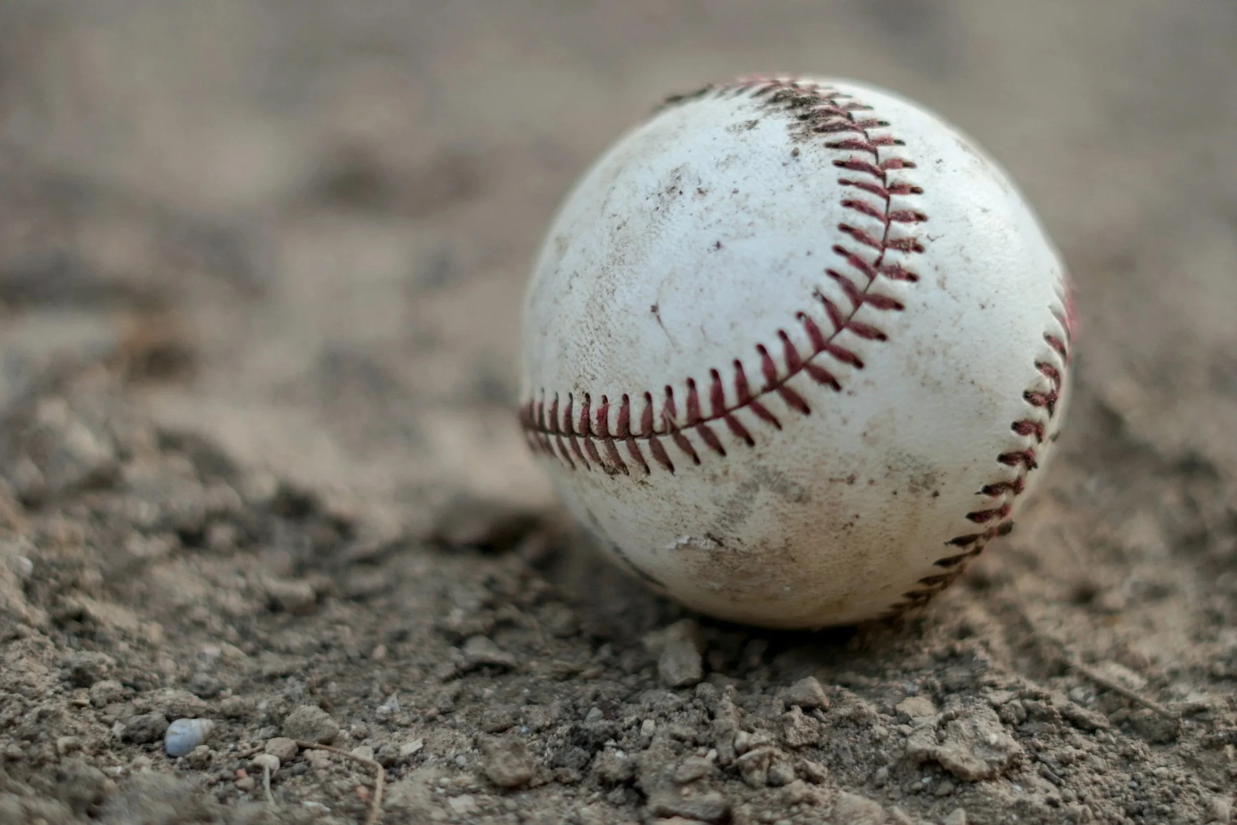 A well-worn baseball with red stitching, resting on dirt ground.
