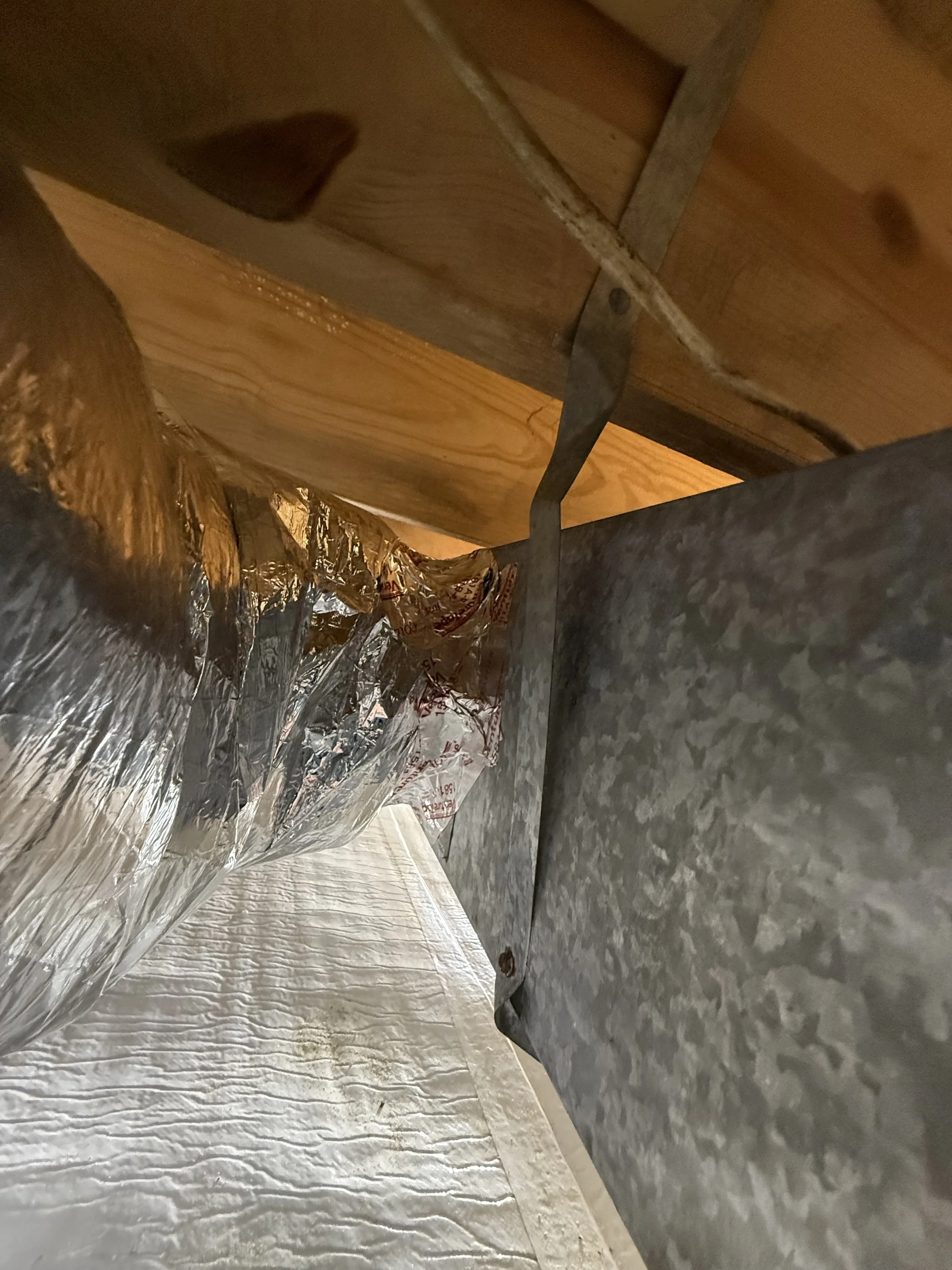 Inside a building, shows a ceiling with wooden beams and metal ductwork, with insulation and vapor barrier visible.